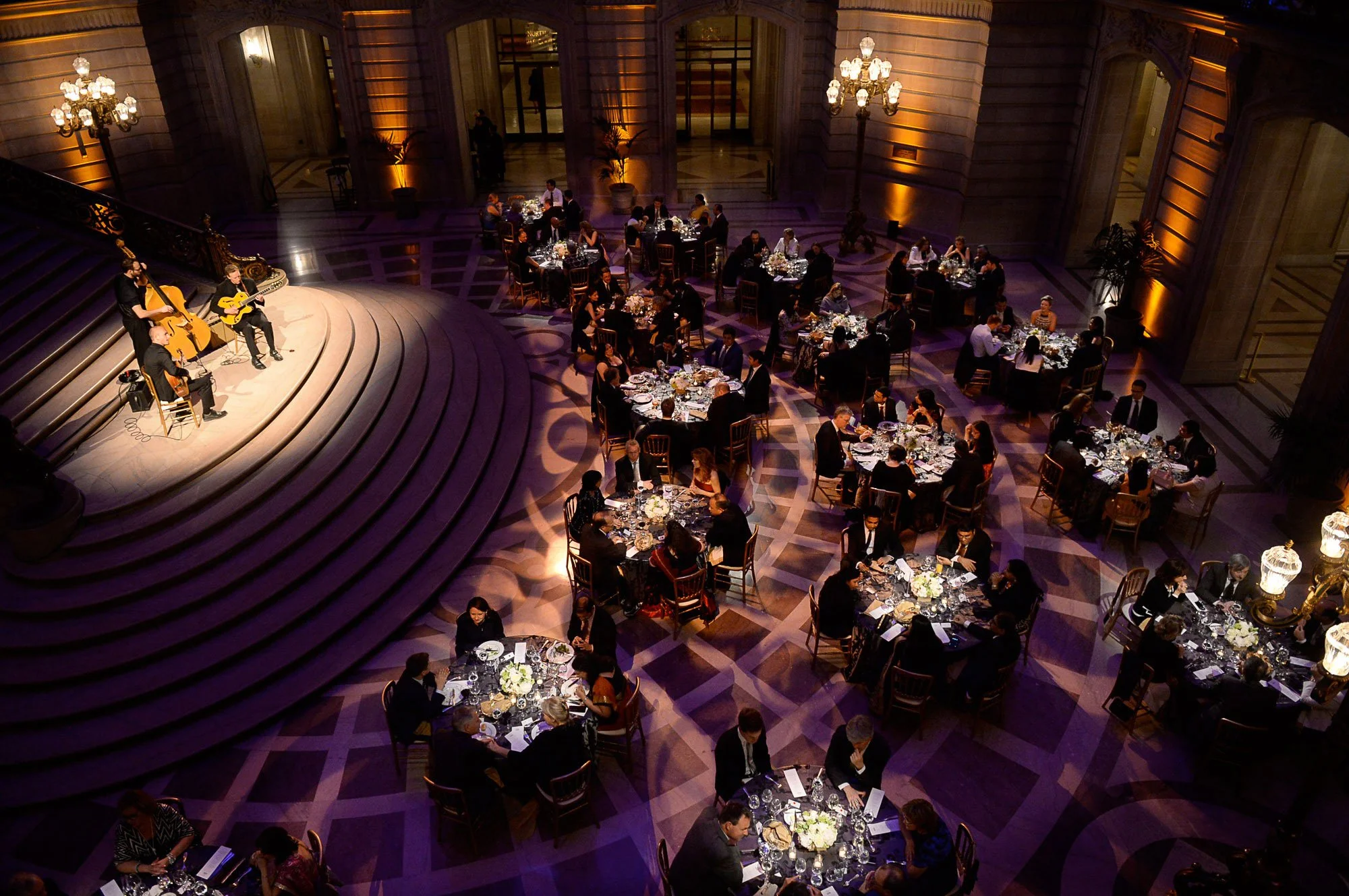 An elegant, large ballroom with high ceilings hosting a formal dinner event. Guests are seated at round tables decorated with floral centerpieces and fine tableware. A small stage on one side features a live musical performance with three musicians p
