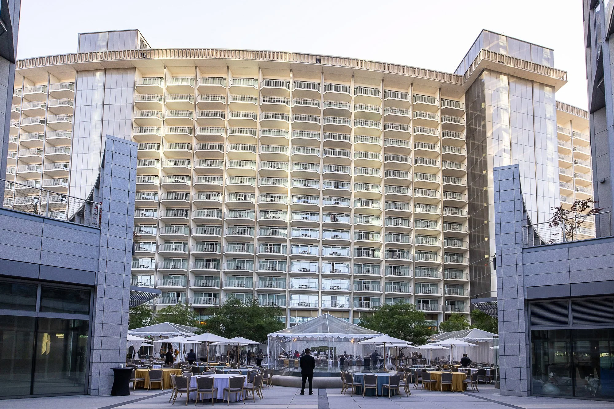 View of an outdoor dining area with tables and chairs, surrounded by tents, in front of a tall, modern hotel building with multiple balconies and large glass windows. The scene is set during the day with clear skies.