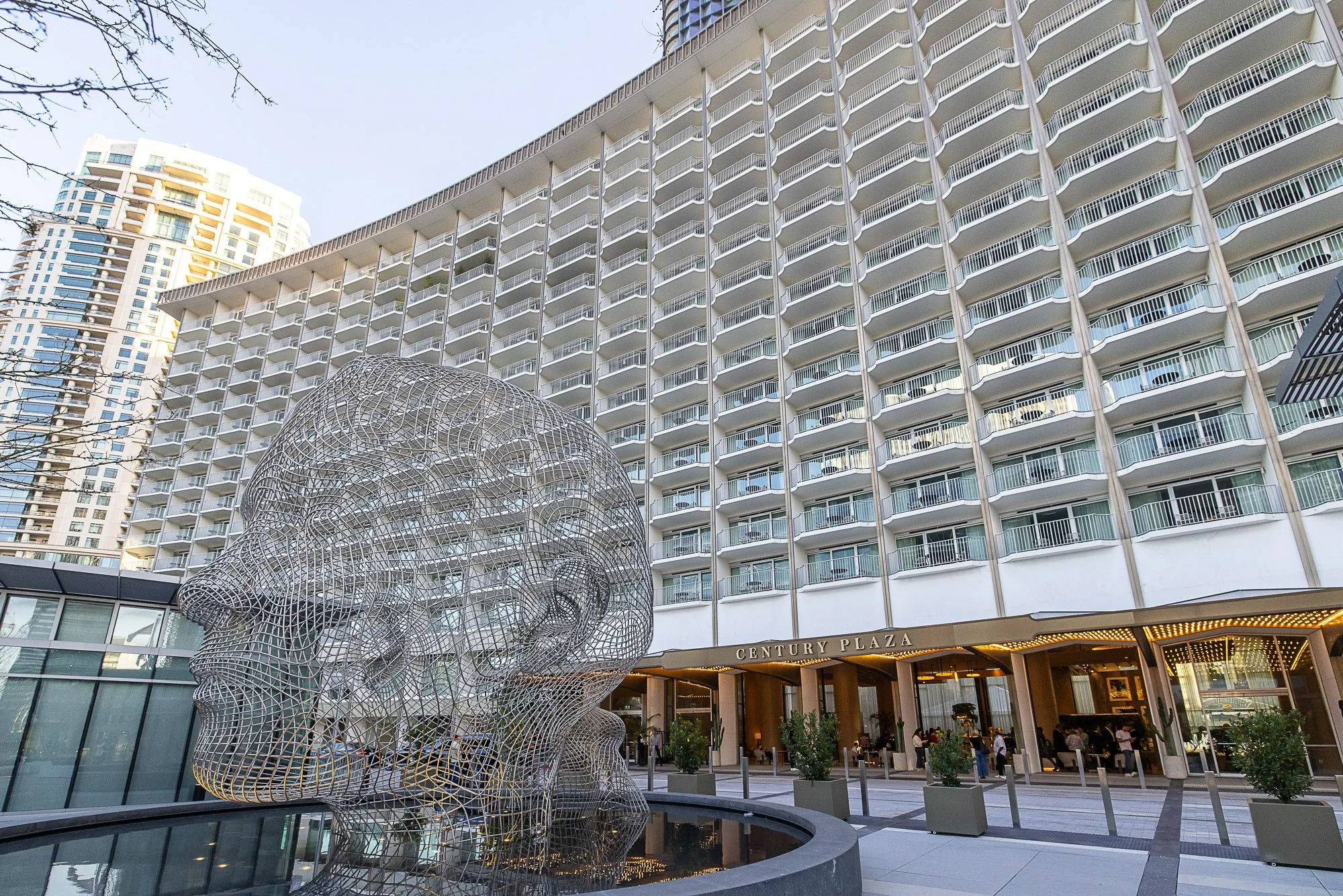 View of a modern high-rise building with balconies, part of Century Plaza, with a distinctive wireframe sculpture of a dog's head in front, and a busy entrance at the bottom with people and potted plants.