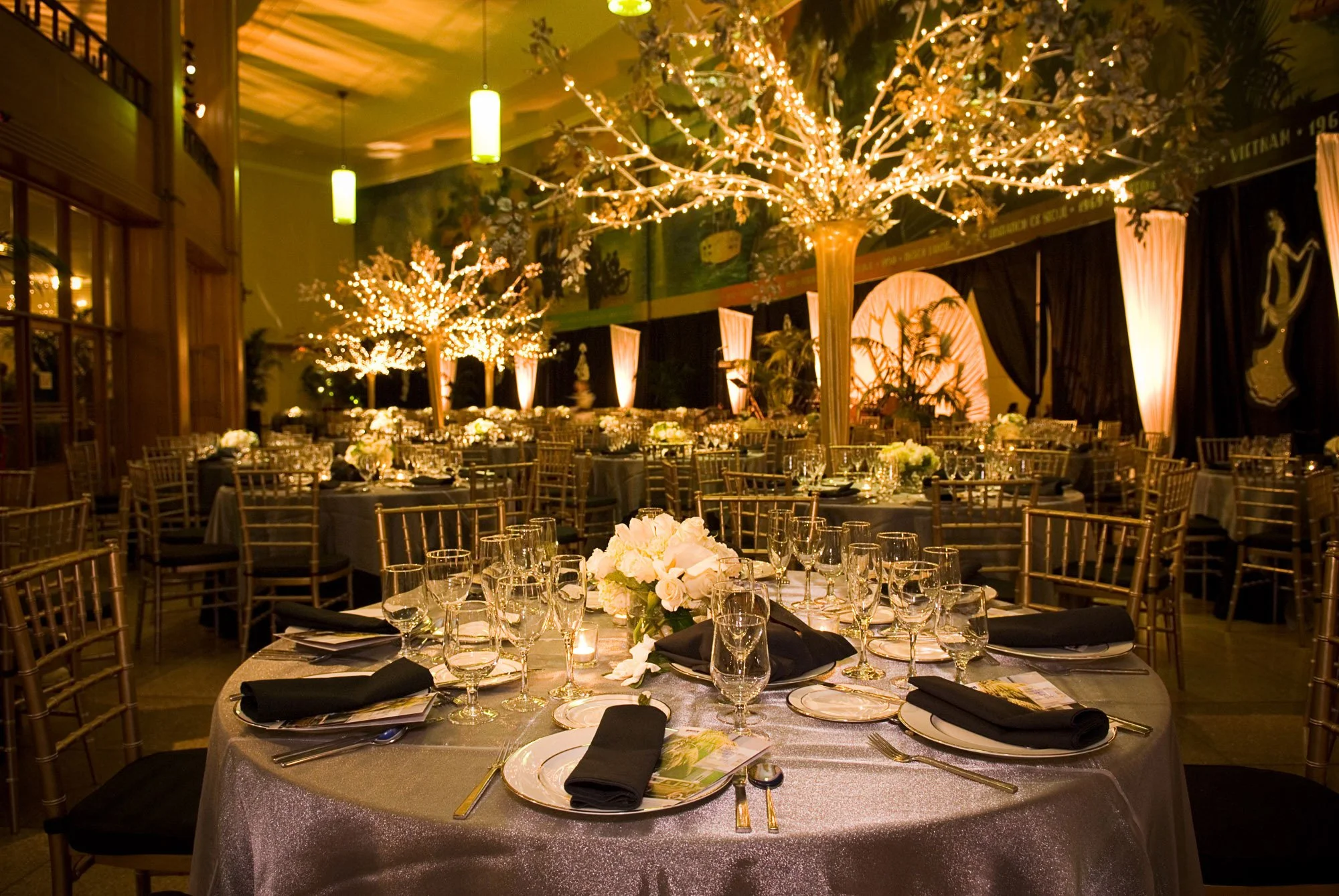 Elegant banquet hall decorated with string lights resembling trees, set up for a formal event with round tables covered with silver tablecloths, floral centerpieces, and place settings including glasses, plates, and black napkins.