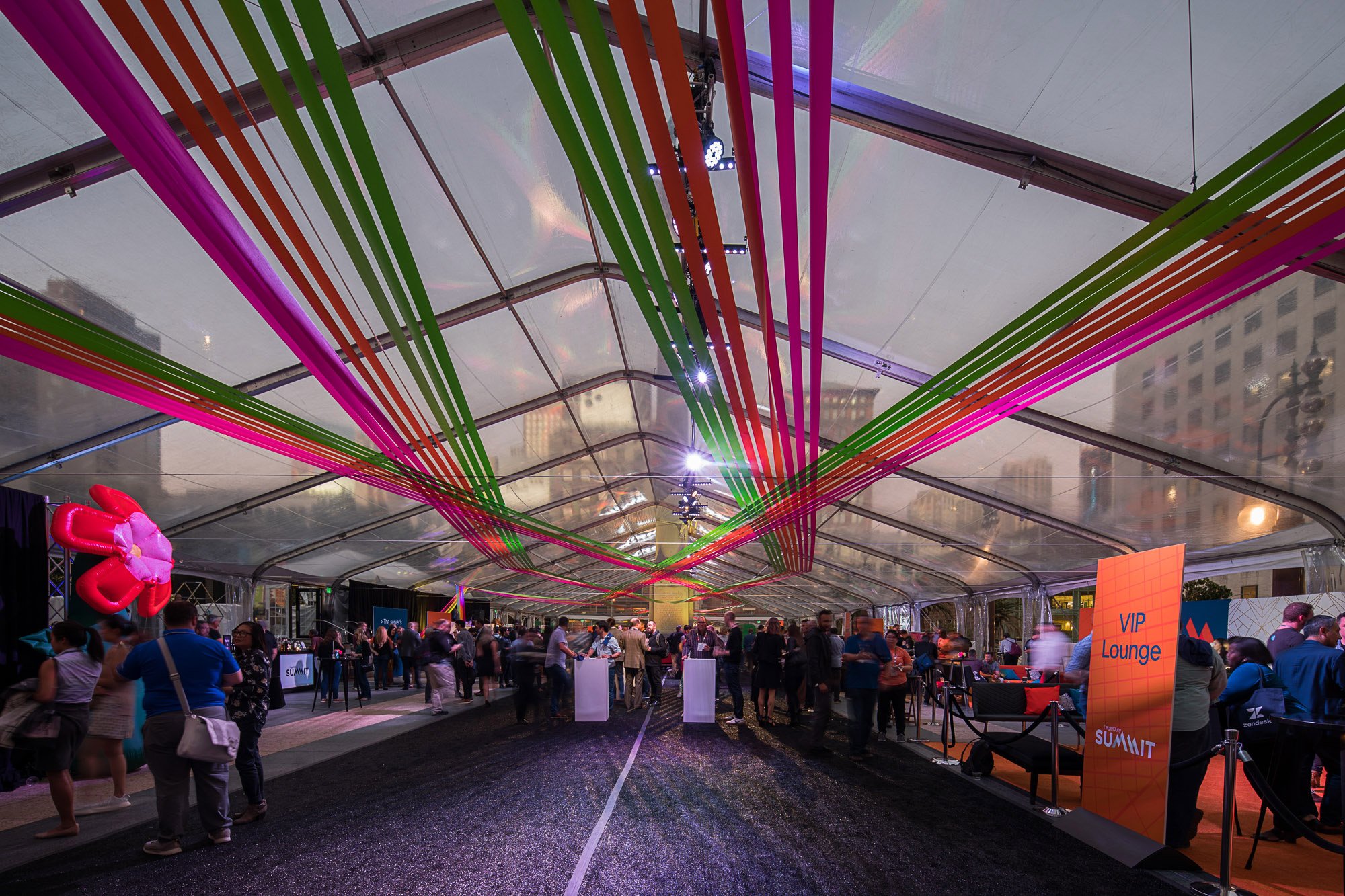 View of a large indoor event with many people under a high, semi-transparent ceiling. Colorful ribbons in pink, green, and orange hang from the ceiling, creating a decorative pattern. There are various booths and signs, including one that reads 'VIP 