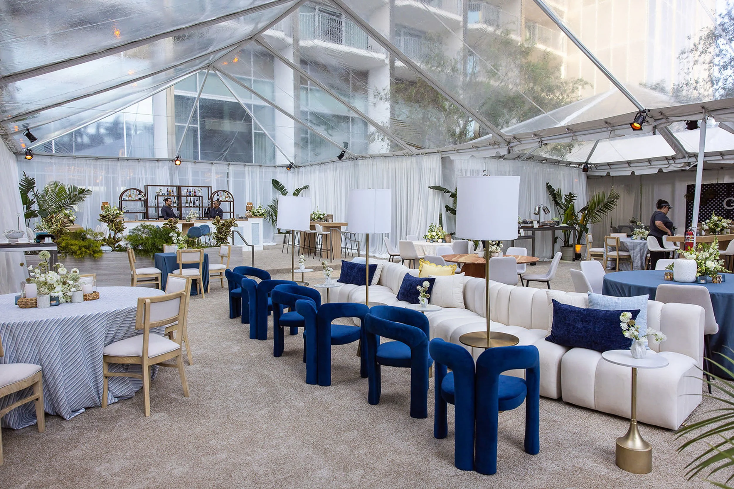 A spacious event tent with a transparent roof, decorated with white, blue, and beige furniture, potted plants, and floral arrangements for a celebration or party.