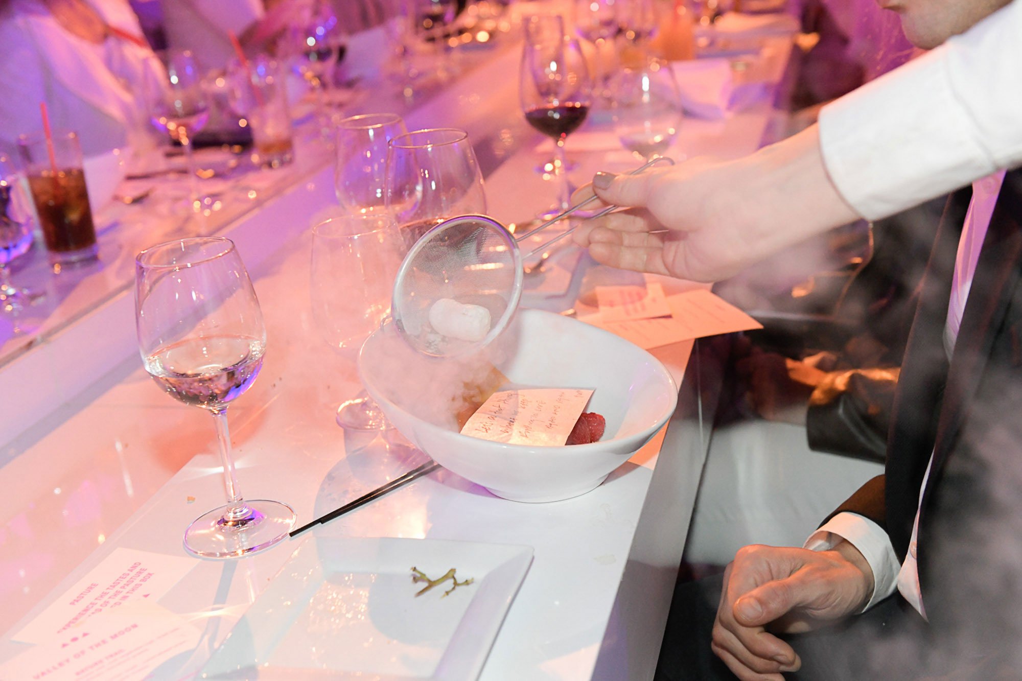 A person in a white shirt is using tongs to place a piece of bread on a plate at a upscale event with a long table set with glasses, wine, and plates.