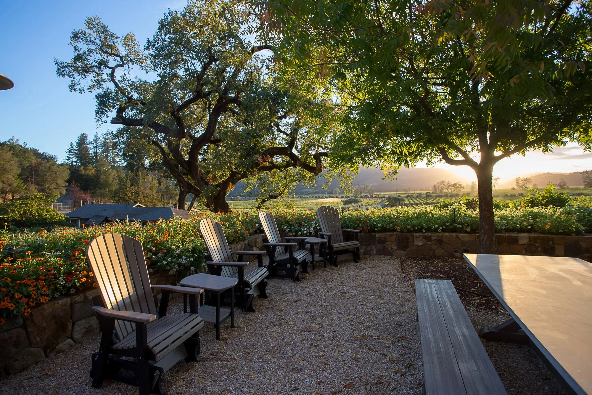 Outdoor patio scene with wooden chairs, a stone wall, a picnic table, surrounded by flowers and trees, with a field and hills in the background during sunset.