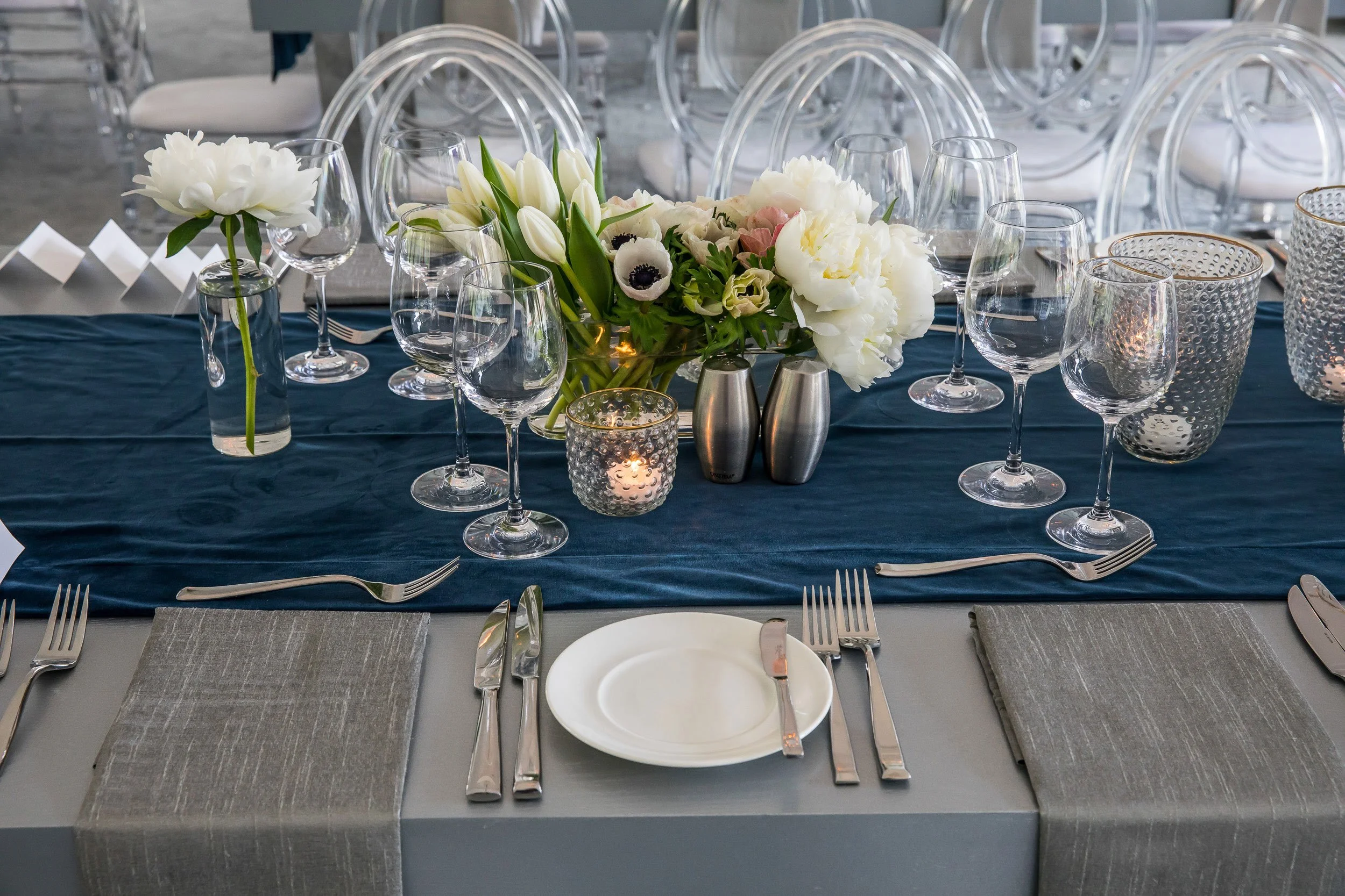 A table set for a formal event with a centerpiece of white flowers and tulips, surrounded by empty wine glasses, silverware, and candles on a dark blue table runner, with transparent chairs in the background.