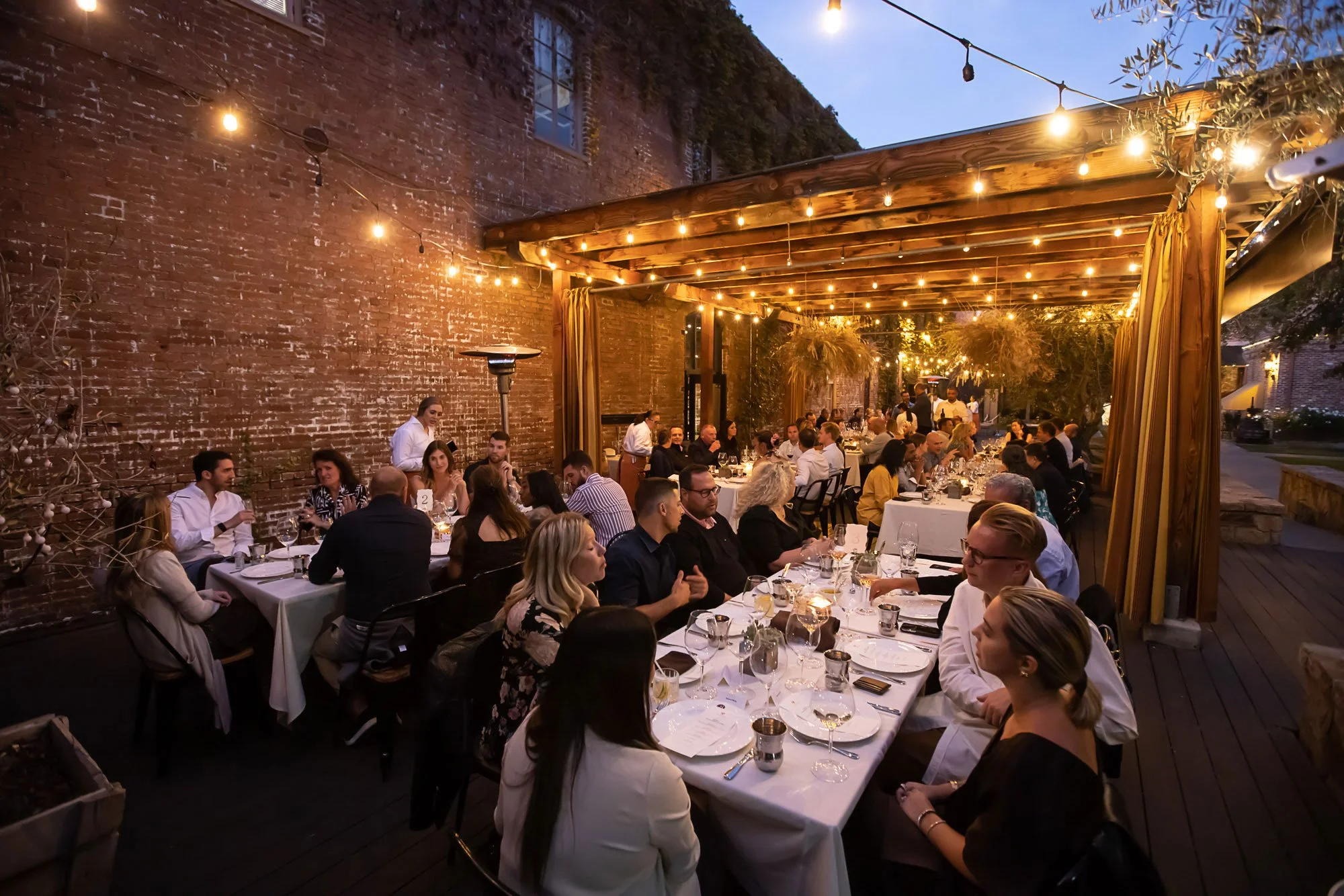 People dining at a long outdoor table under string lights during the evening, with brick walls and trees visible in the background.