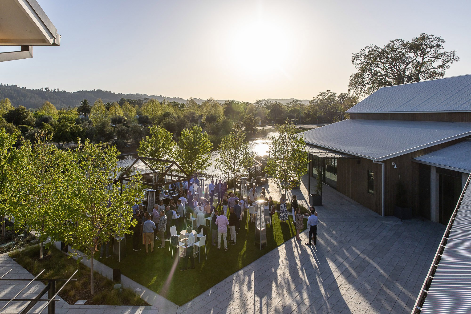 A group of people at an outdoor event on a sunny day, with trees, a river, and hills in the background, near modern buildings with metal roofs.