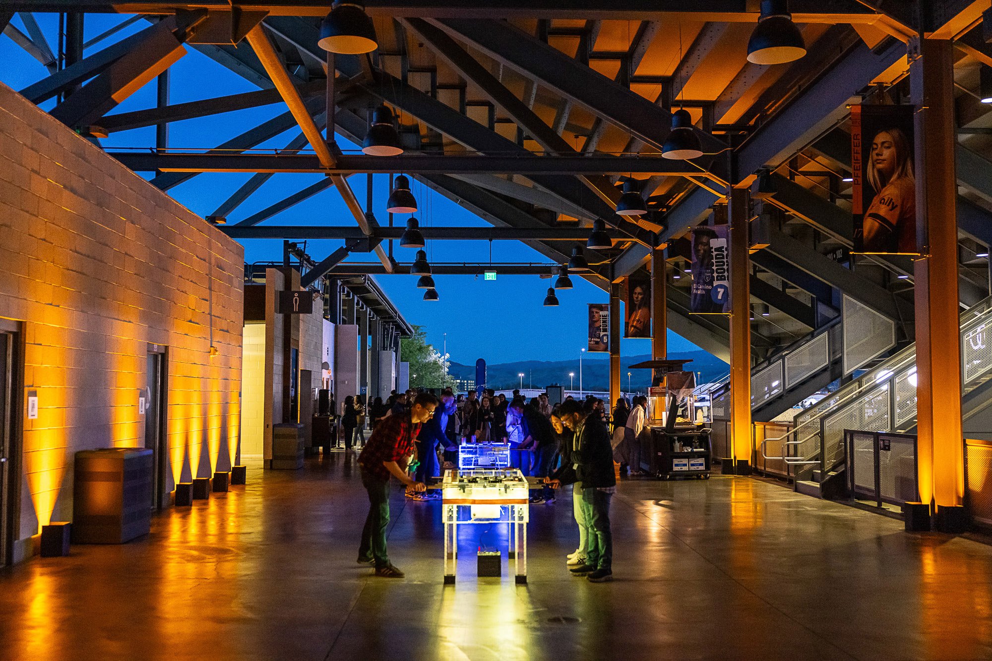 A crowded indoor space with people gathered around illuminated tables, under a large open ceiling with metal beams and industrial-style lighting, during the evening or night.