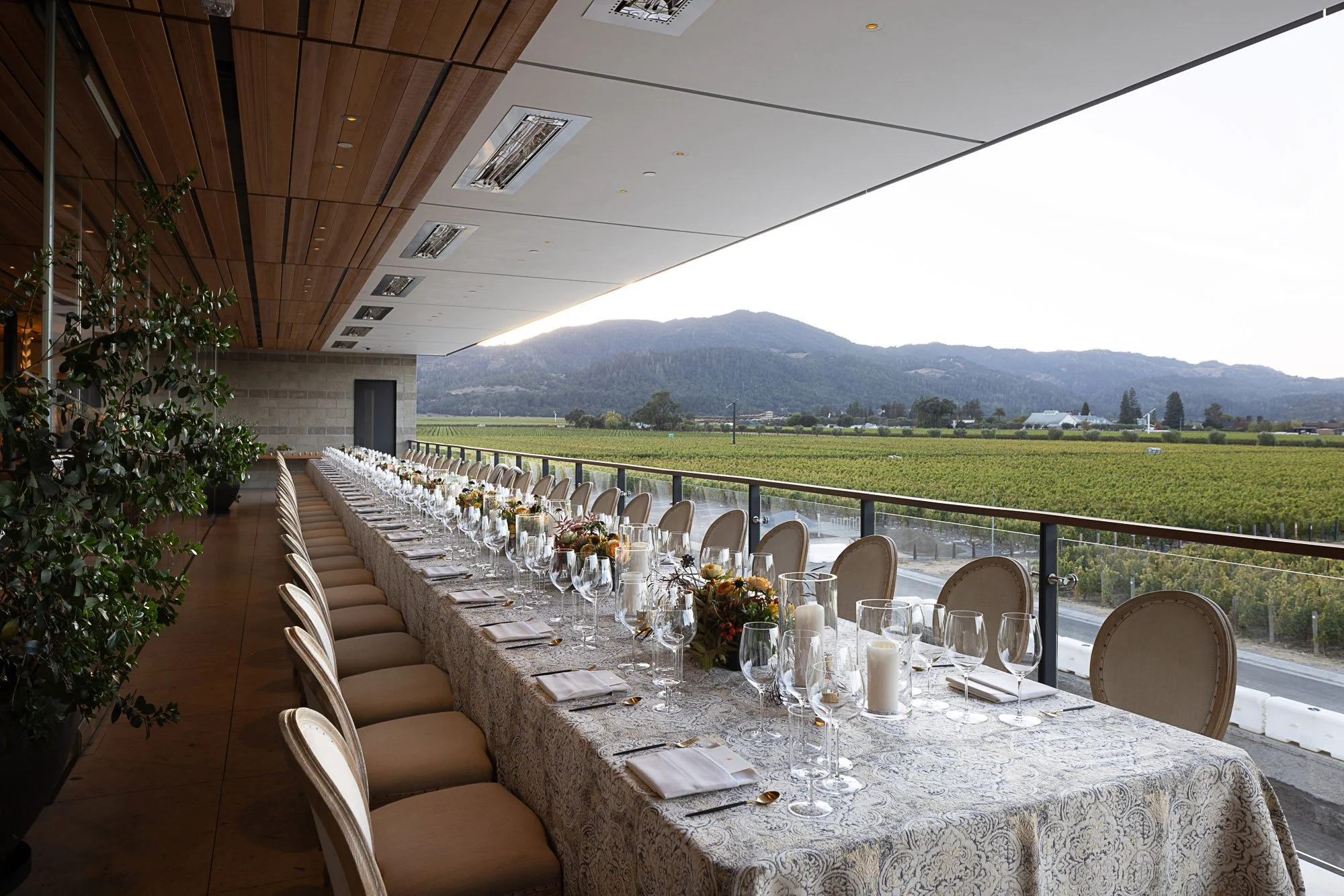 Long dining table set for a formal event with chairs, glassware, and floral centerpieces on a decorated table, indoors overlooking agricultural fields and mountains.