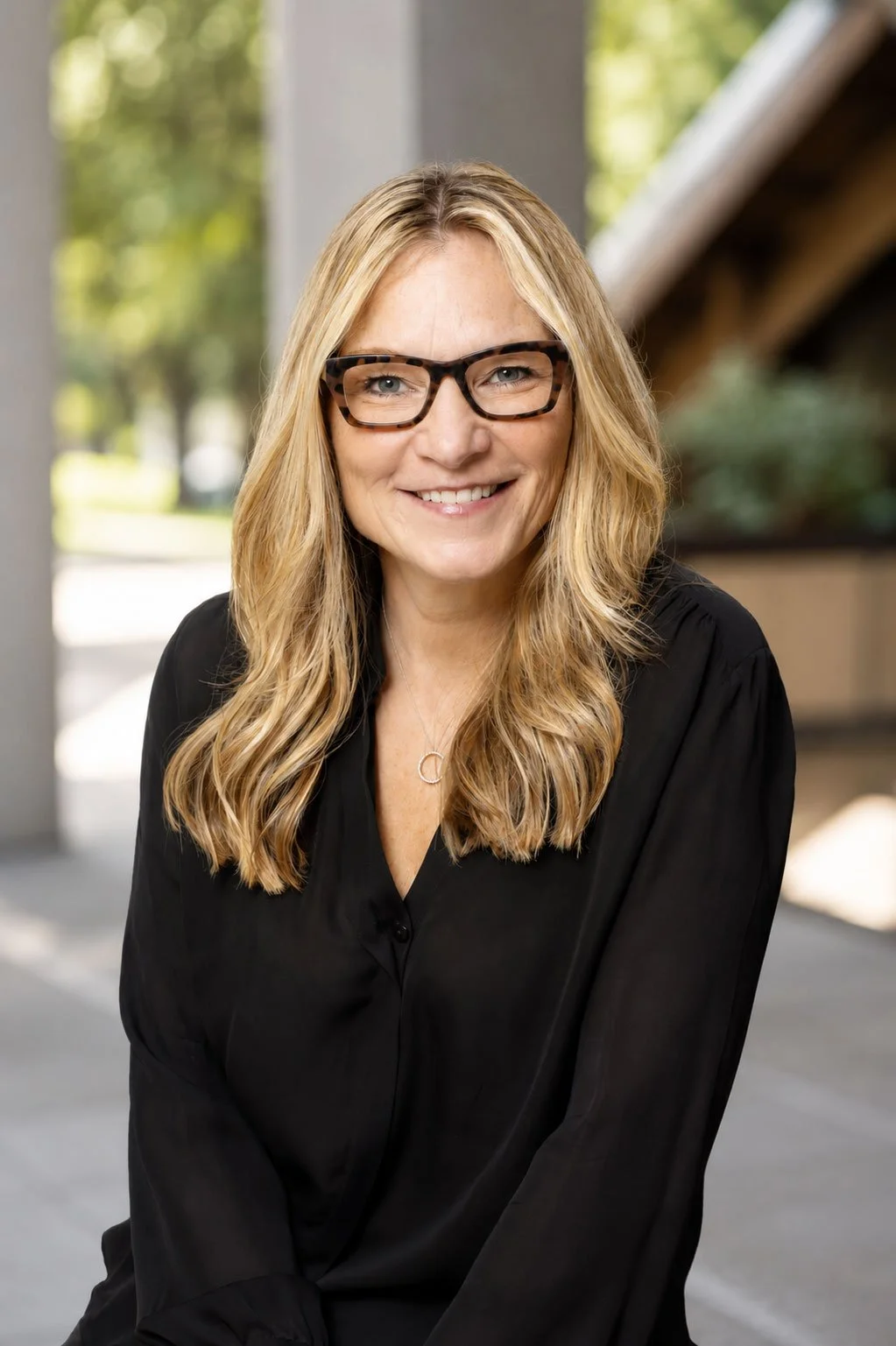 A woman with wavy blonde hair, wearing glasses and a black top, smiling at the camera outdoors with trees and a building structure in the background.
