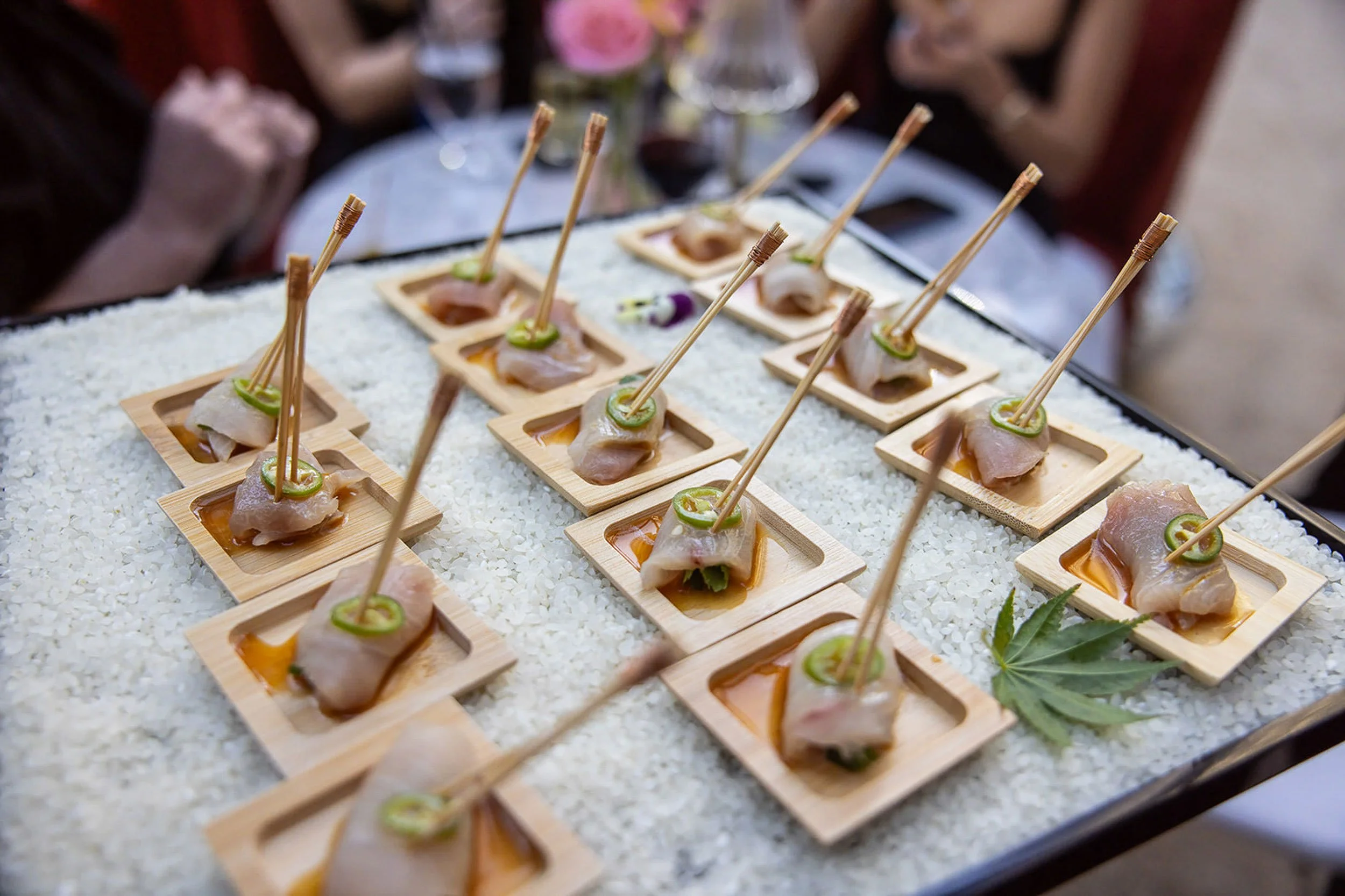 Close-up of elegant appetizers arranged on small wooden plates, featuring sliced meat with a slice of jalapeño chili, set on a bed of white rice with a green leaf garnish, at a formal dining event.
