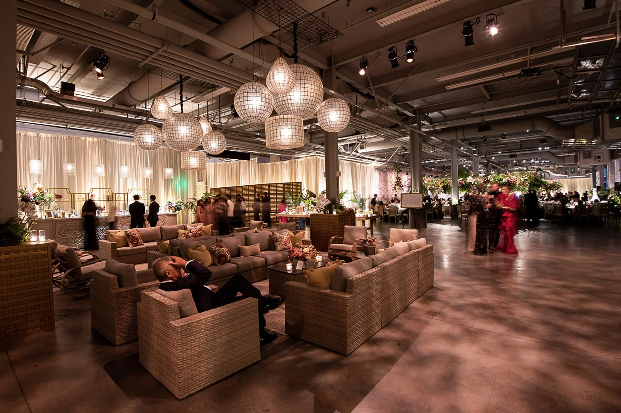 Interior view of a decorated event space with hanging pendant lights, draped curtains, floral arrangements, and guests socializing and seated on wicker furniture.