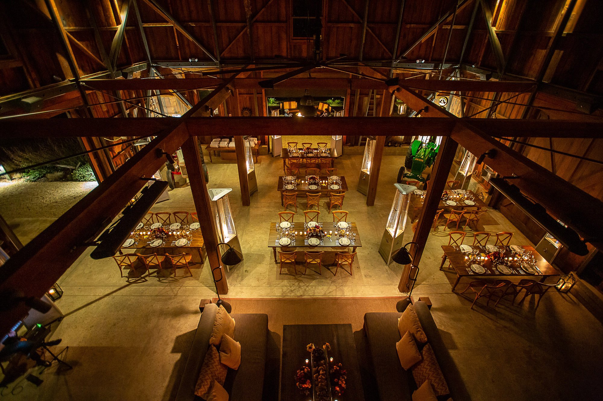 An interior view of a rustic dining room with wooden beams and tables set for a meal, decorated with wine glasses and candles, with chairs around the tables, warm lighting, and a cozy atmosphere.