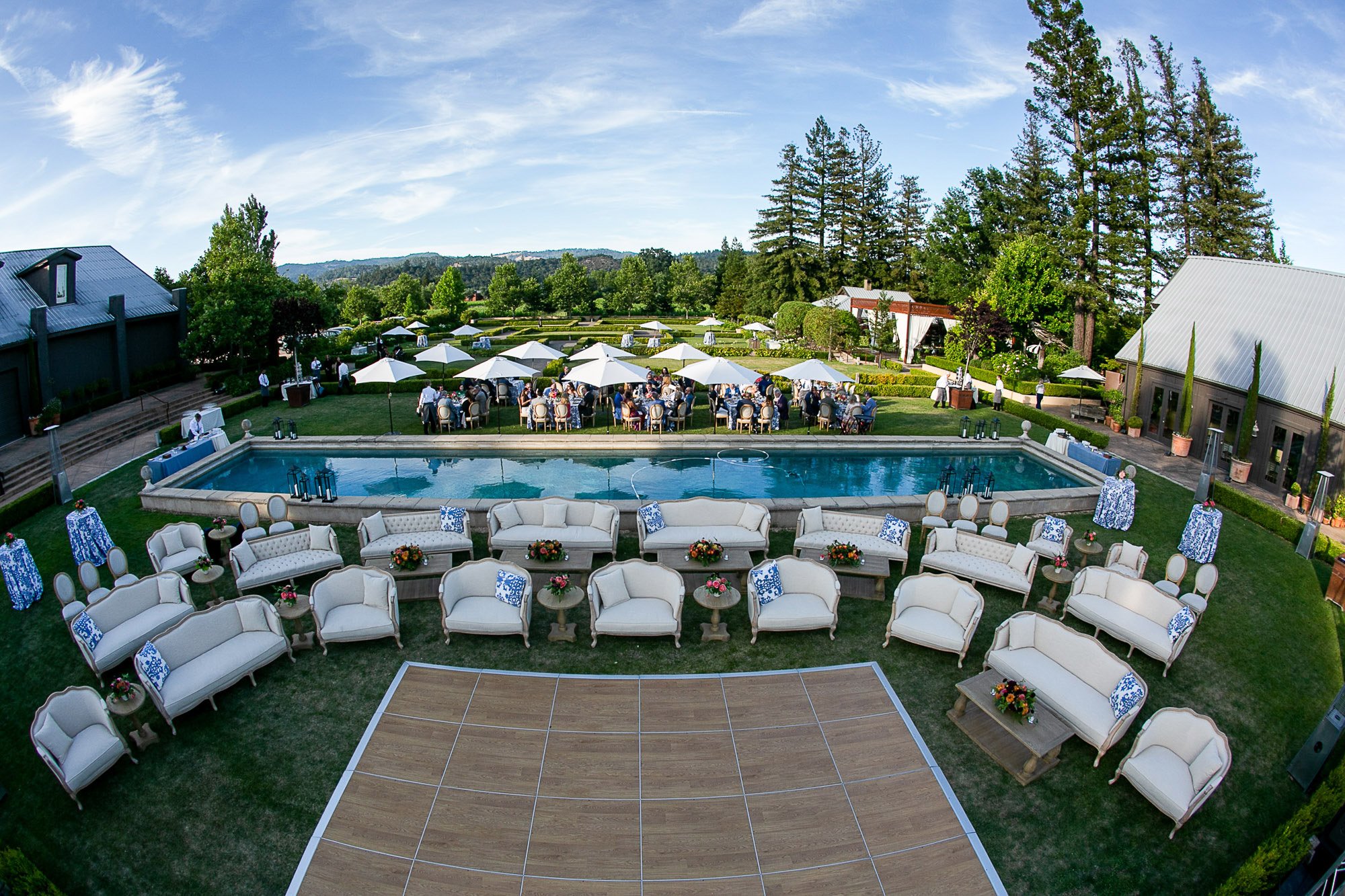 Outdoor wedding reception setup with elegant white and blue decorated furniture near a swimming pool, white umbrellas, lush green trees, and a scenic landscape in the background.