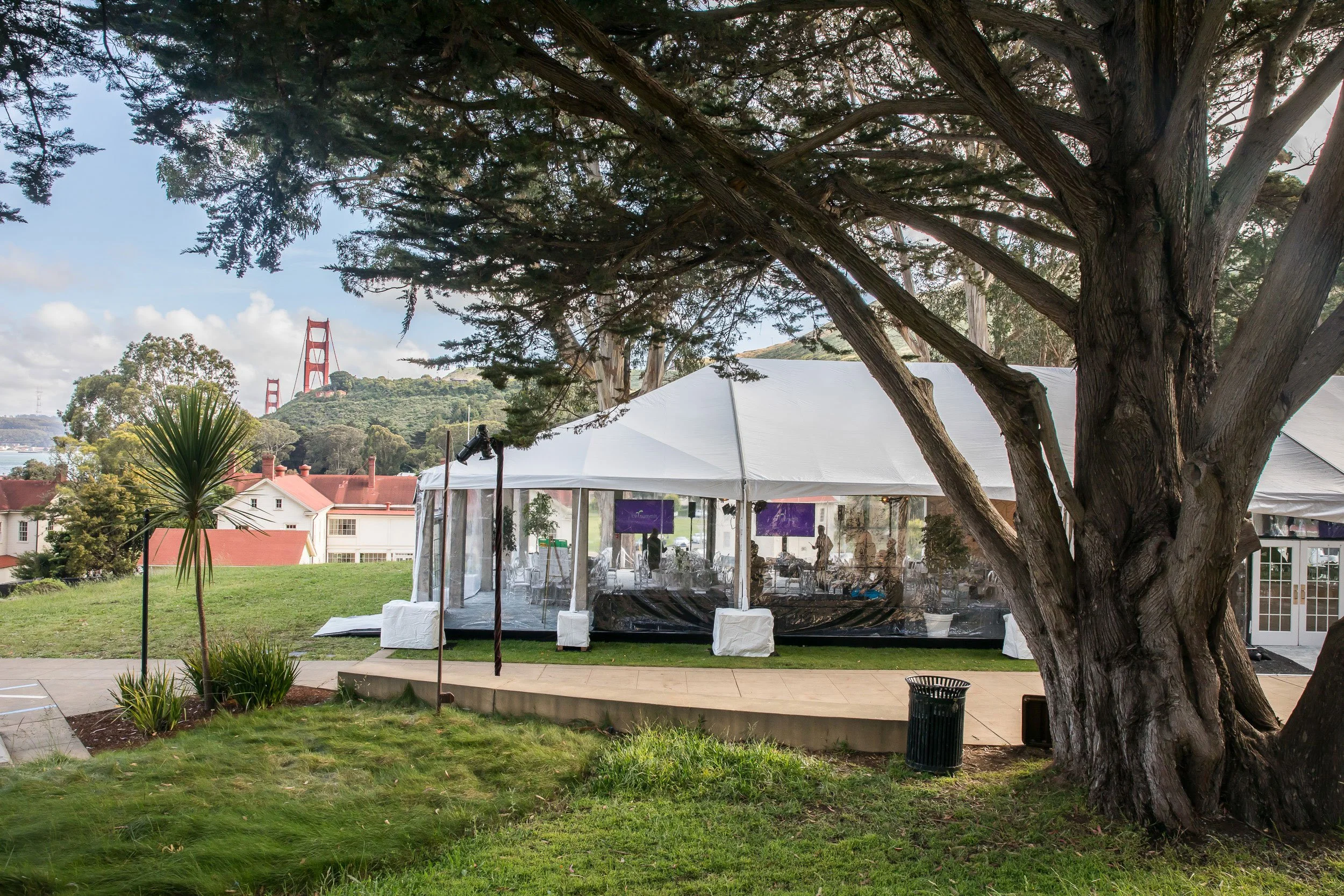 An outdoor event setup with a large white tent, trees, and a view of the Golden Gate Bridge in the background on a partly cloudy day.