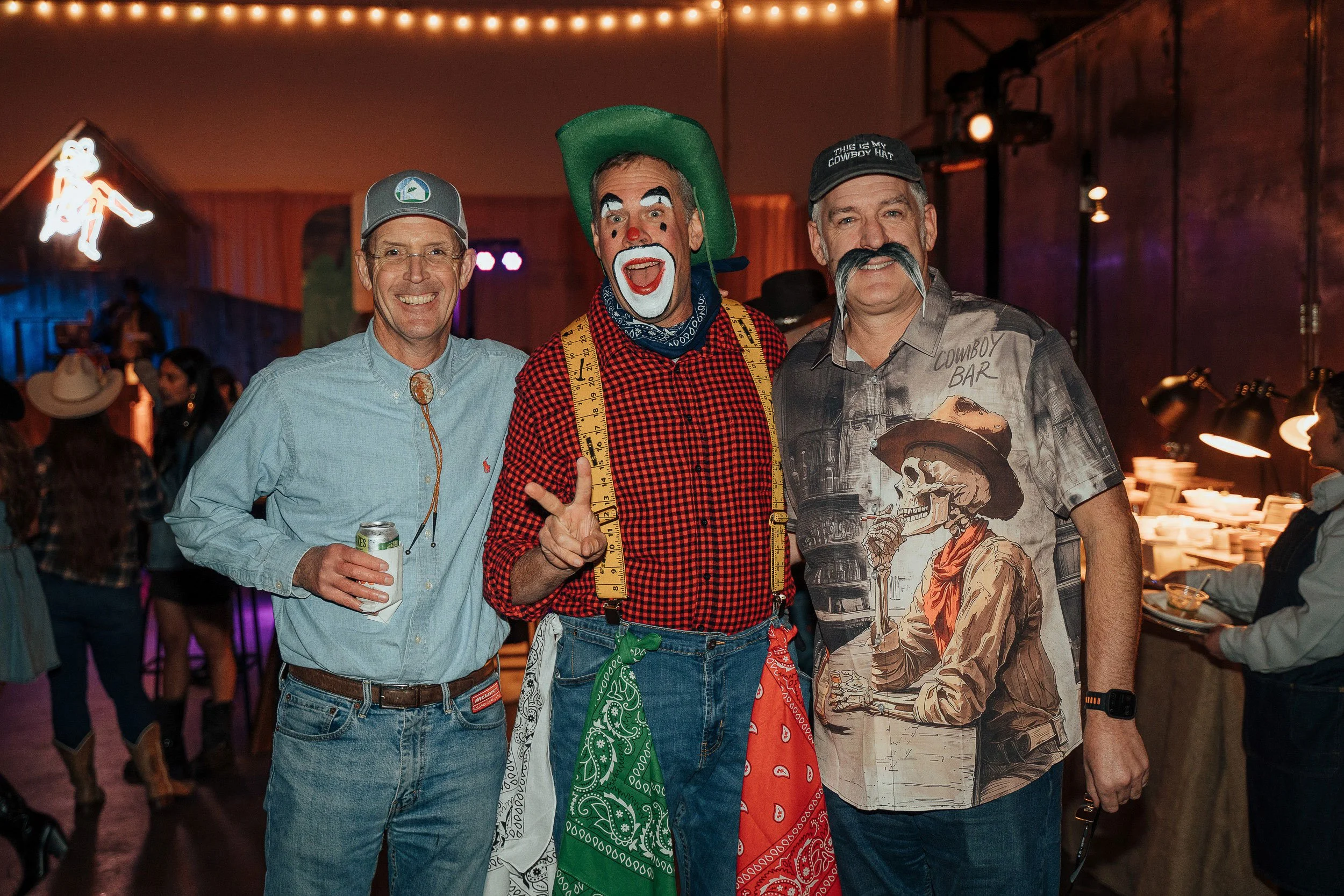 Three men smiling at a party, with a clown in the middle wearing a green hat, red checkered shirt, and a measuring tape around his neck, while the man on the right is wearing a cowboy-themed shirt and a baseball cap.