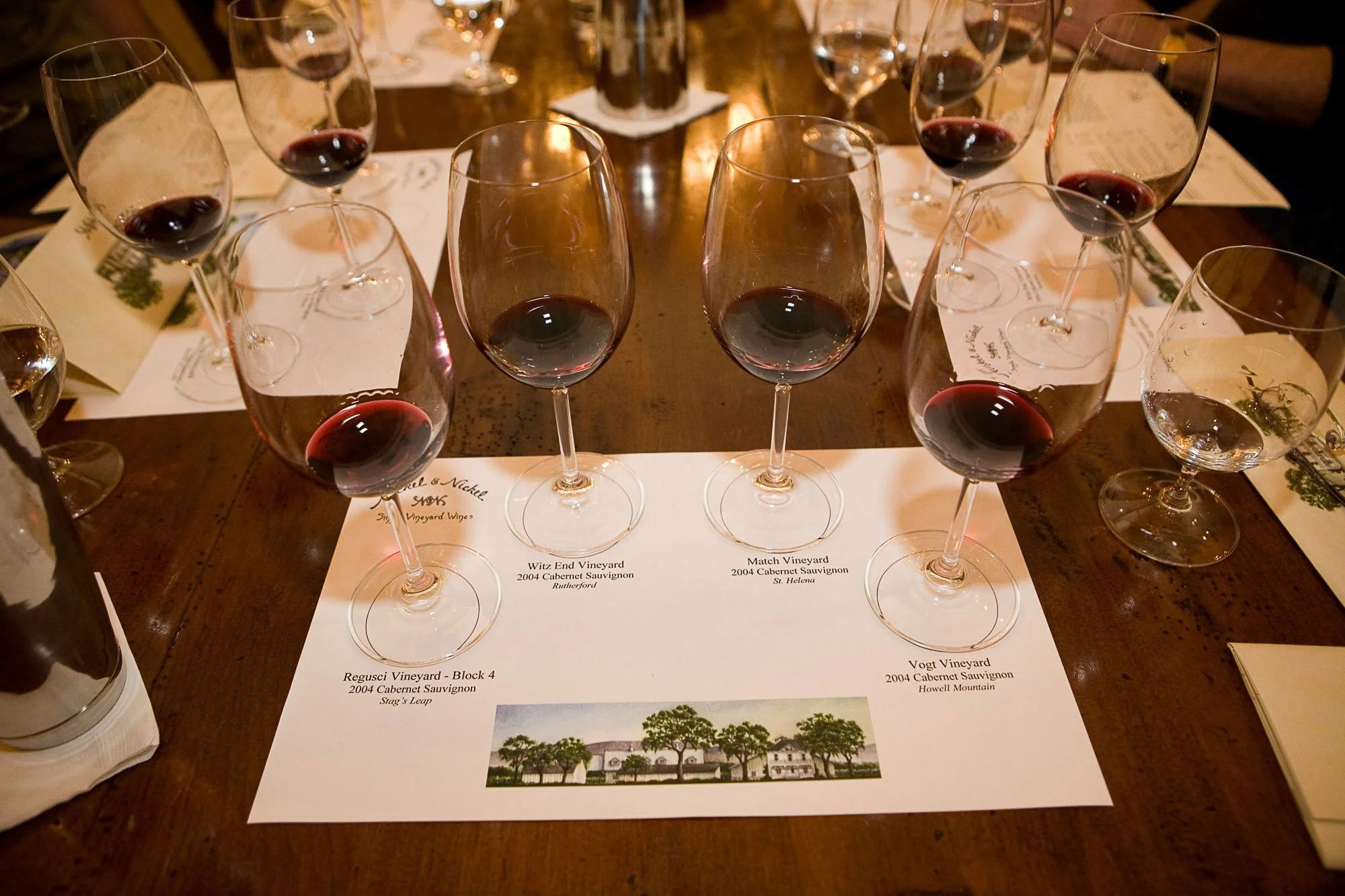 A table with multiple wine glasses filled with red wine, placed over sheets of paper with wine descriptions and images of vineyards.