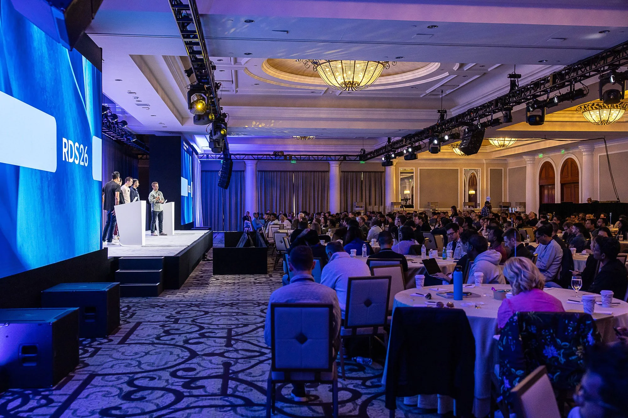A conference room filled with attendees sitting at round tables, facing a stage with a large blue screen displaying 'RDS26' and four speakers standing at a podium. The room features chandeliers and professional lighting equipment overhead.