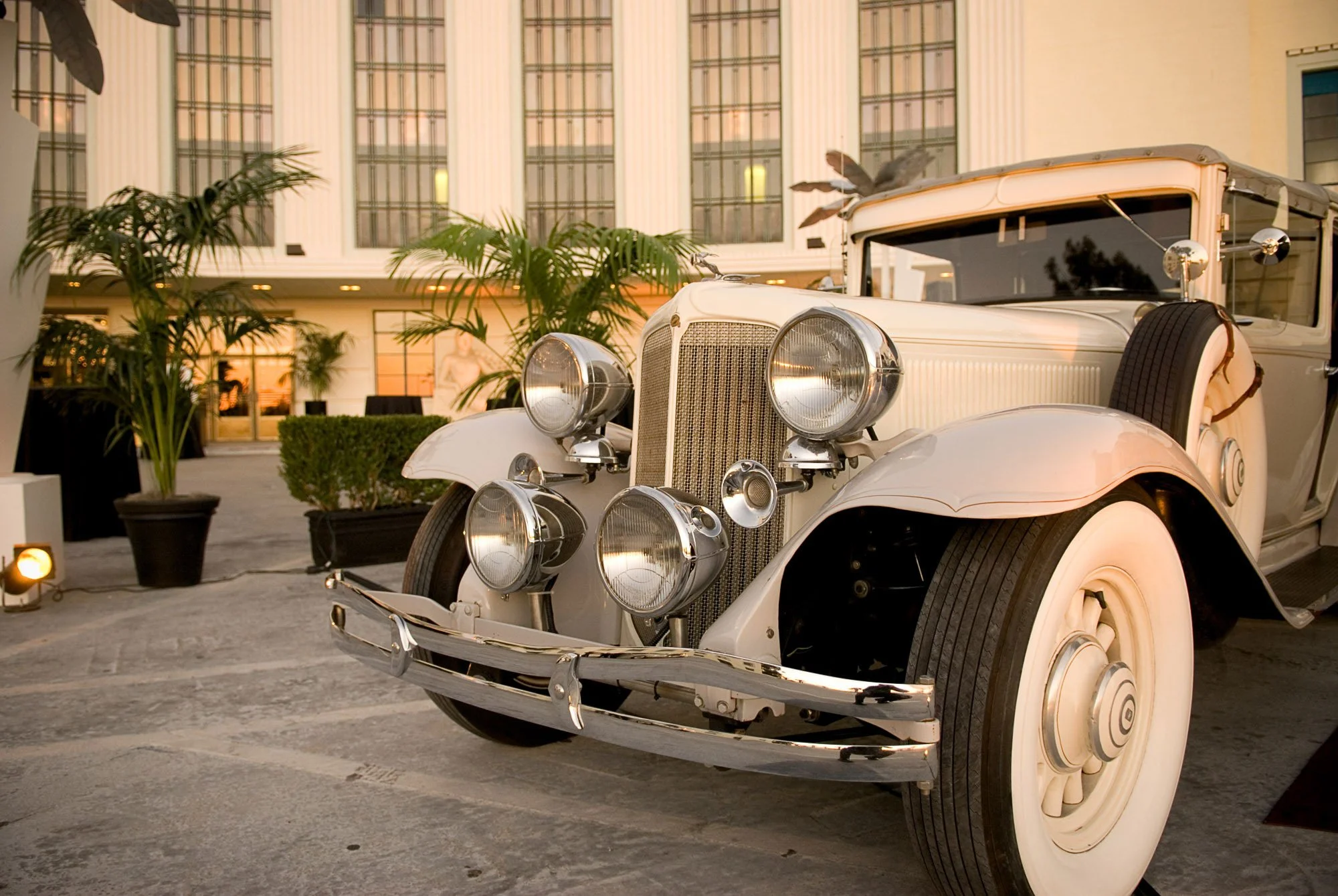 Vintage white car with large round headlights and chrome details parked outdoors near a modern building with large windows and columns. Potted plants are visible nearby, with warm lighting.