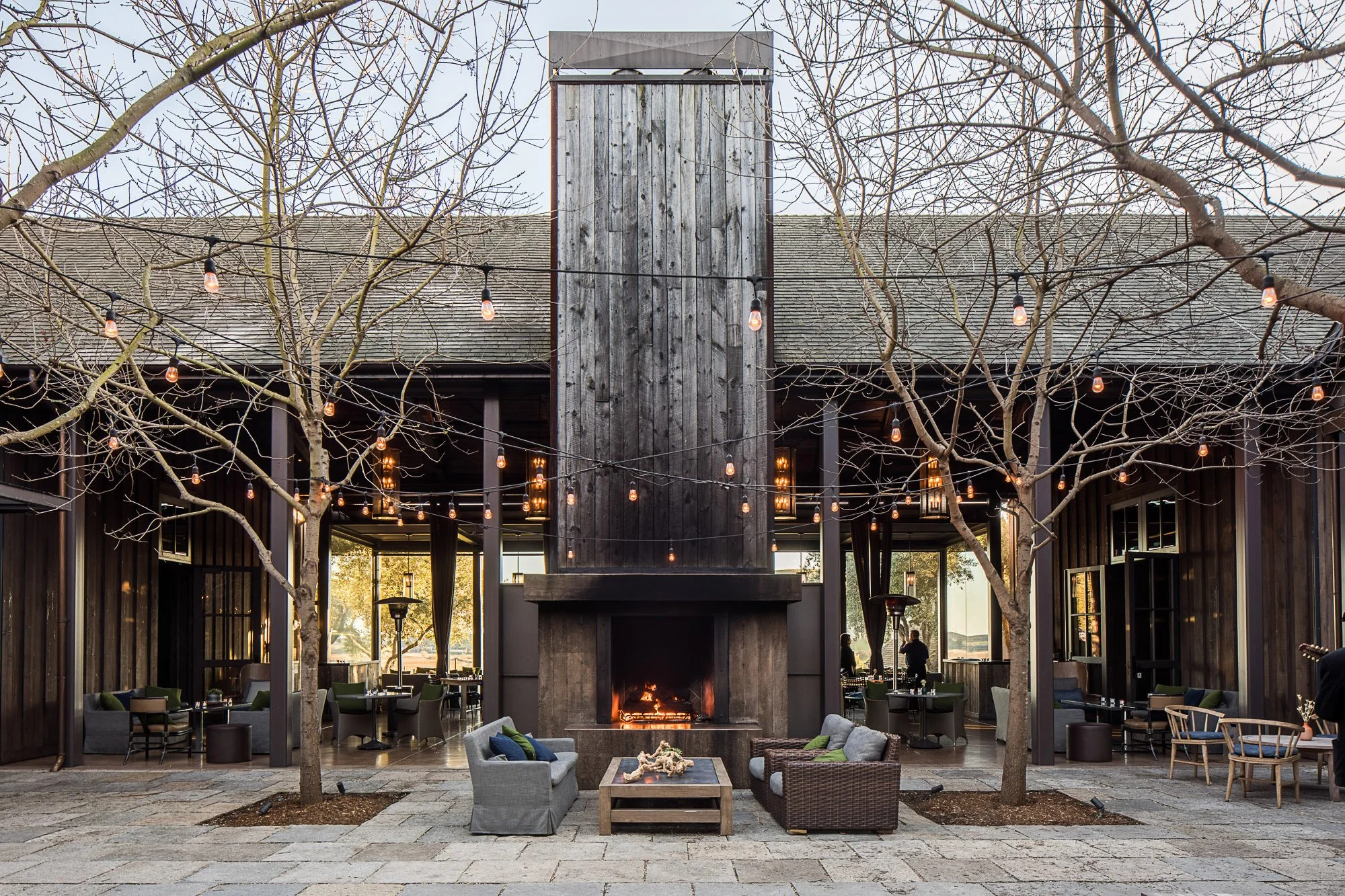 An outdoor seating area features a large central fireplace with a black wooden chimney, surrounded by tables, chairs, and sofas. String lights hang across the area, and leafless trees frame the scene. The background shows a building with large window