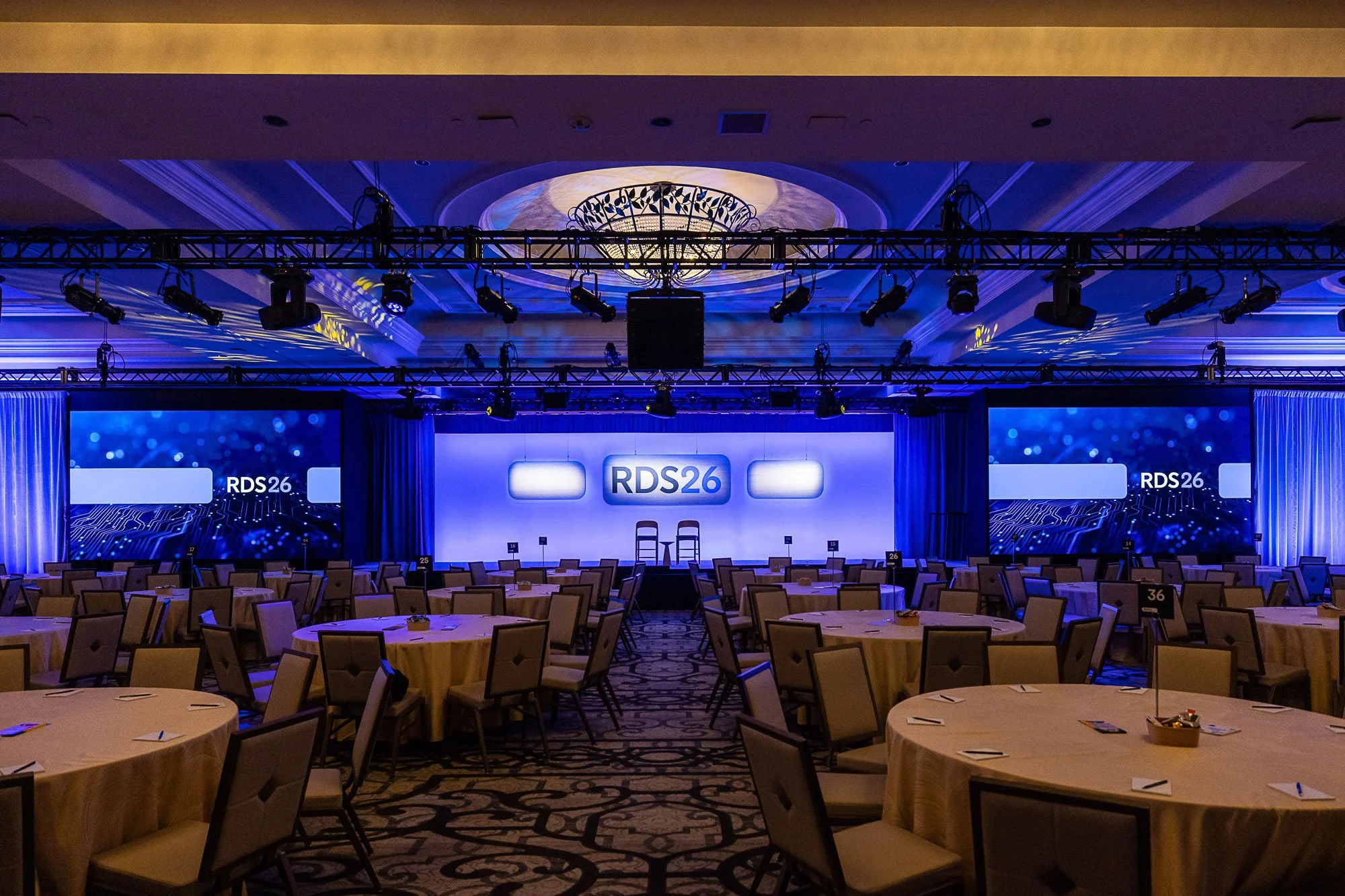 A large conference room with round tables covered in beige tablecloths, each with chairs around them. The stage has a large screen displaying 'RDS26' and is equipped with lighting and audio equipment. The room is decorated in a professional manner su