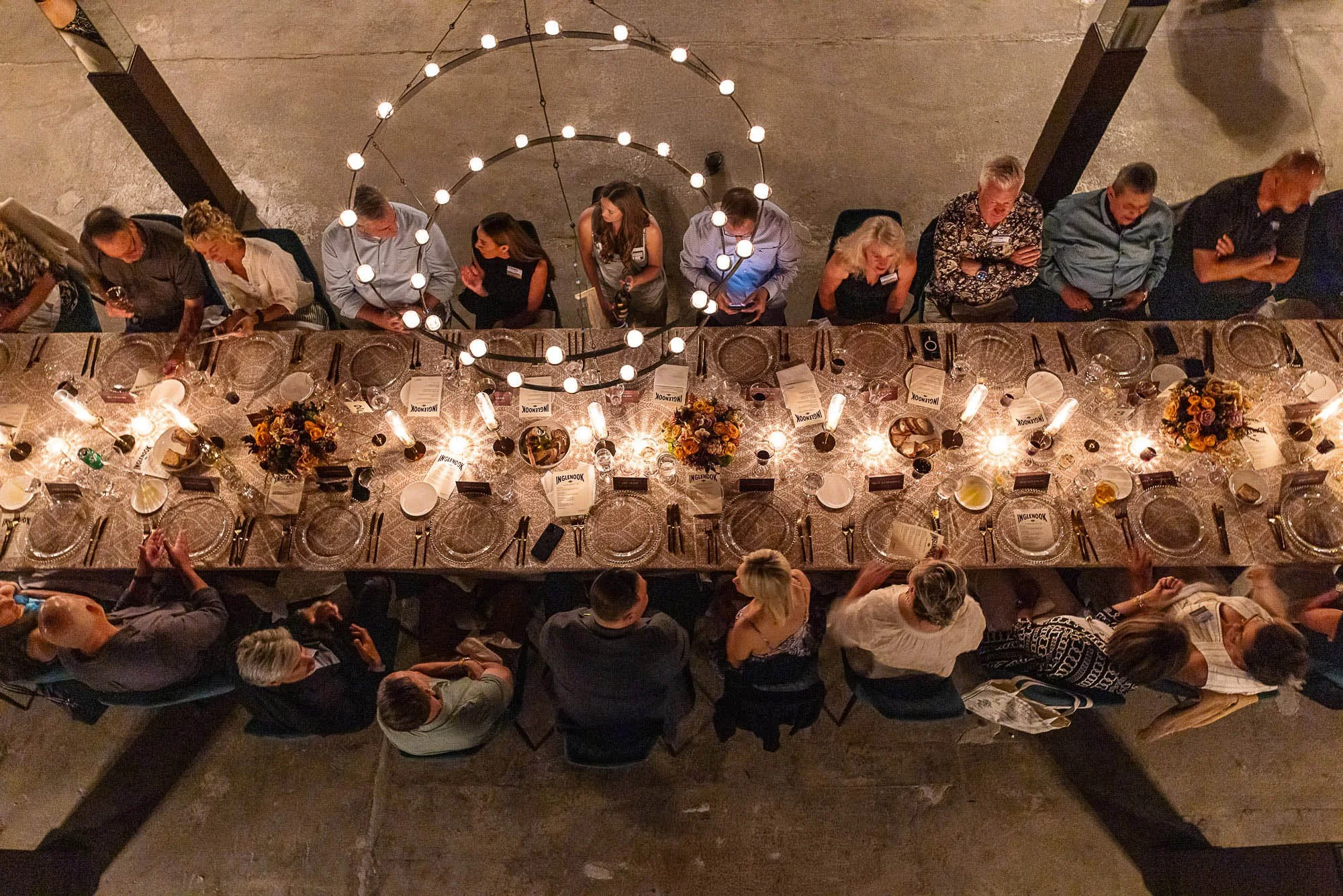 Overhead view of a long banquet table set for a meal with floral centerpieces, wine glasses, and tableware, with people standing and seated around it, in a warmly lit room.