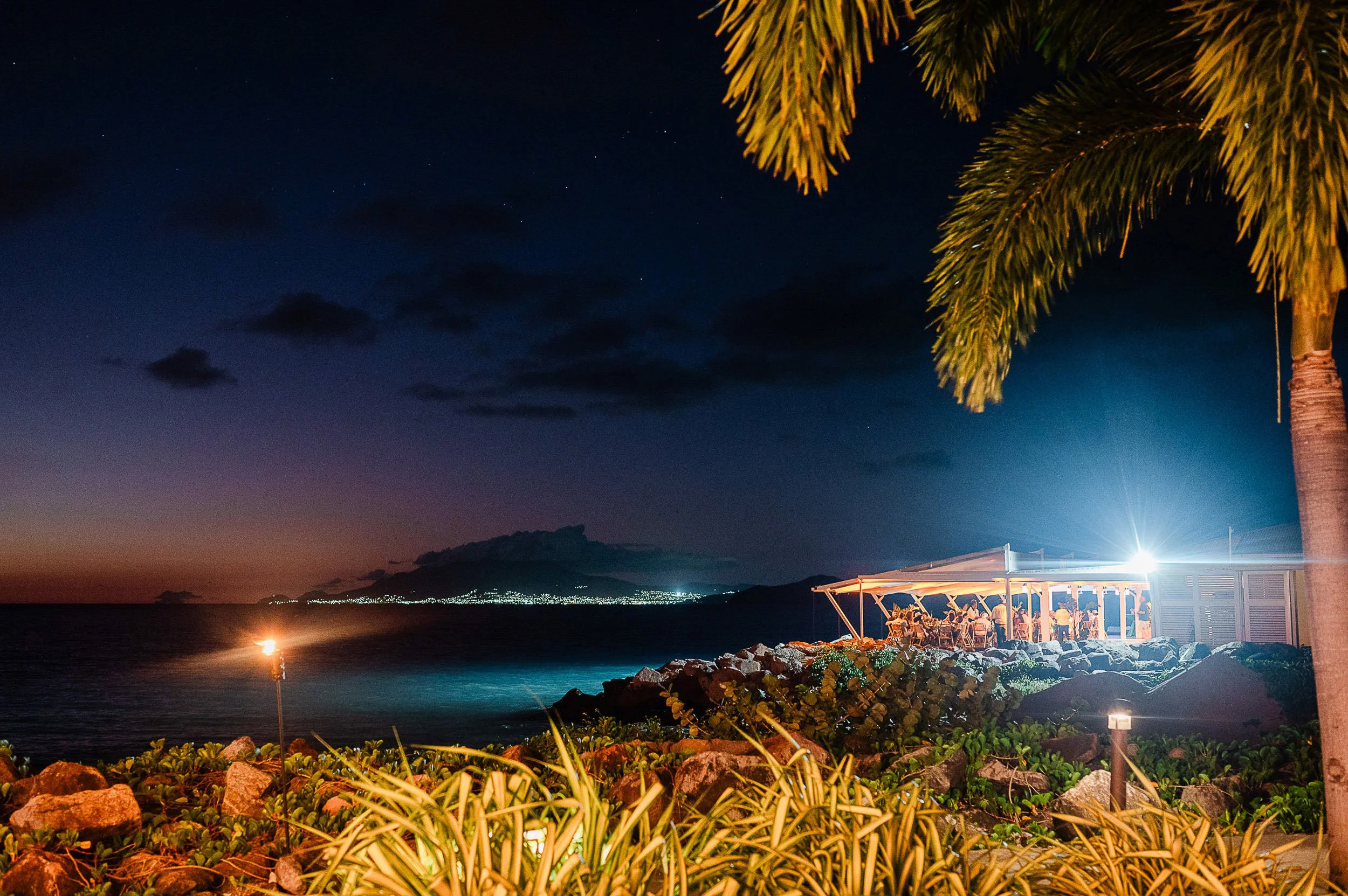 Night scene of a beachside restaurant with tables and chairs, palm trees in the foreground, and a clear starry sky with a few clouds in the background.