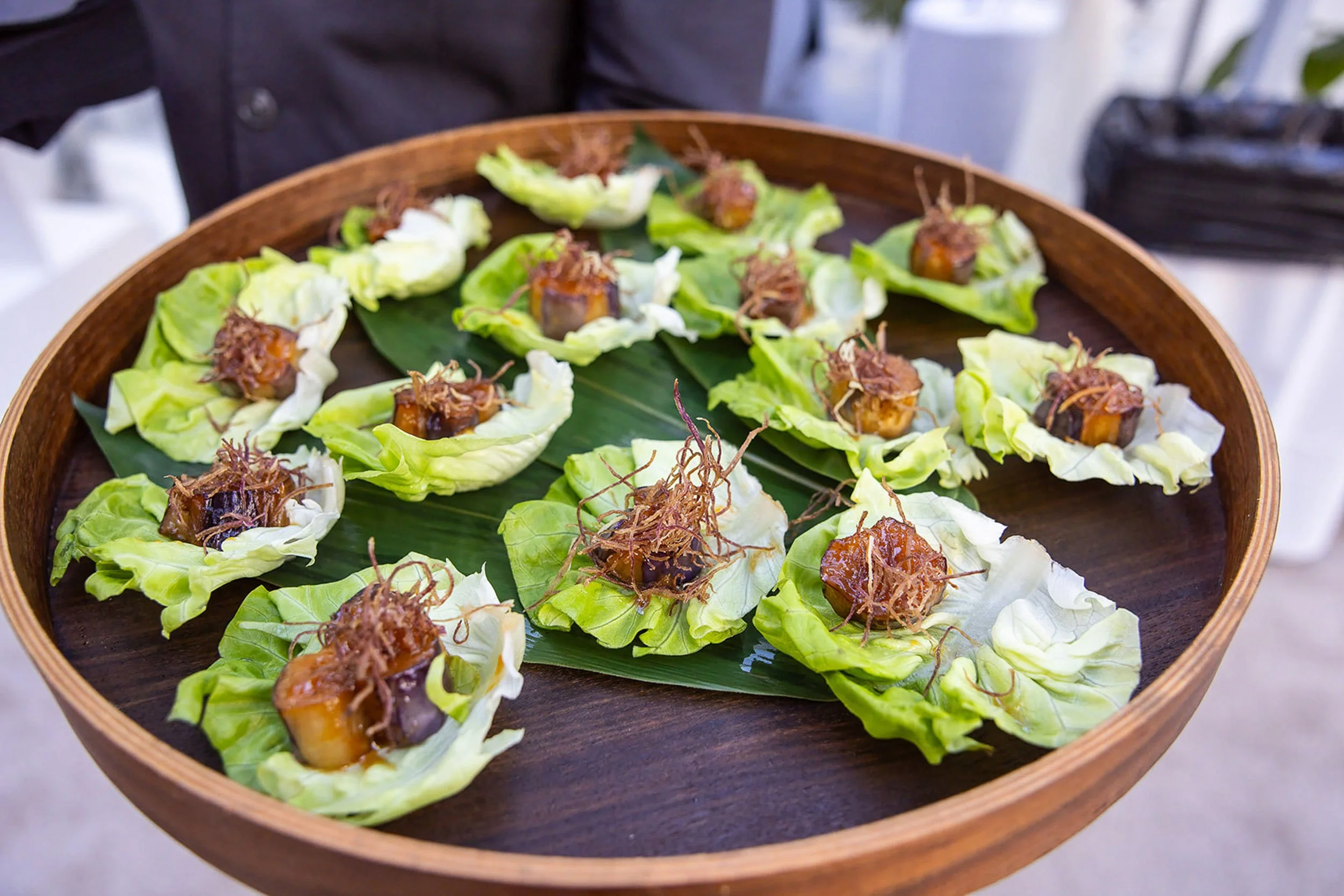 Small appetizers served on lettuce leaves with a topping and garnished with crispy fried strands, arranged on a round wooden tray lined with a large green leaf.