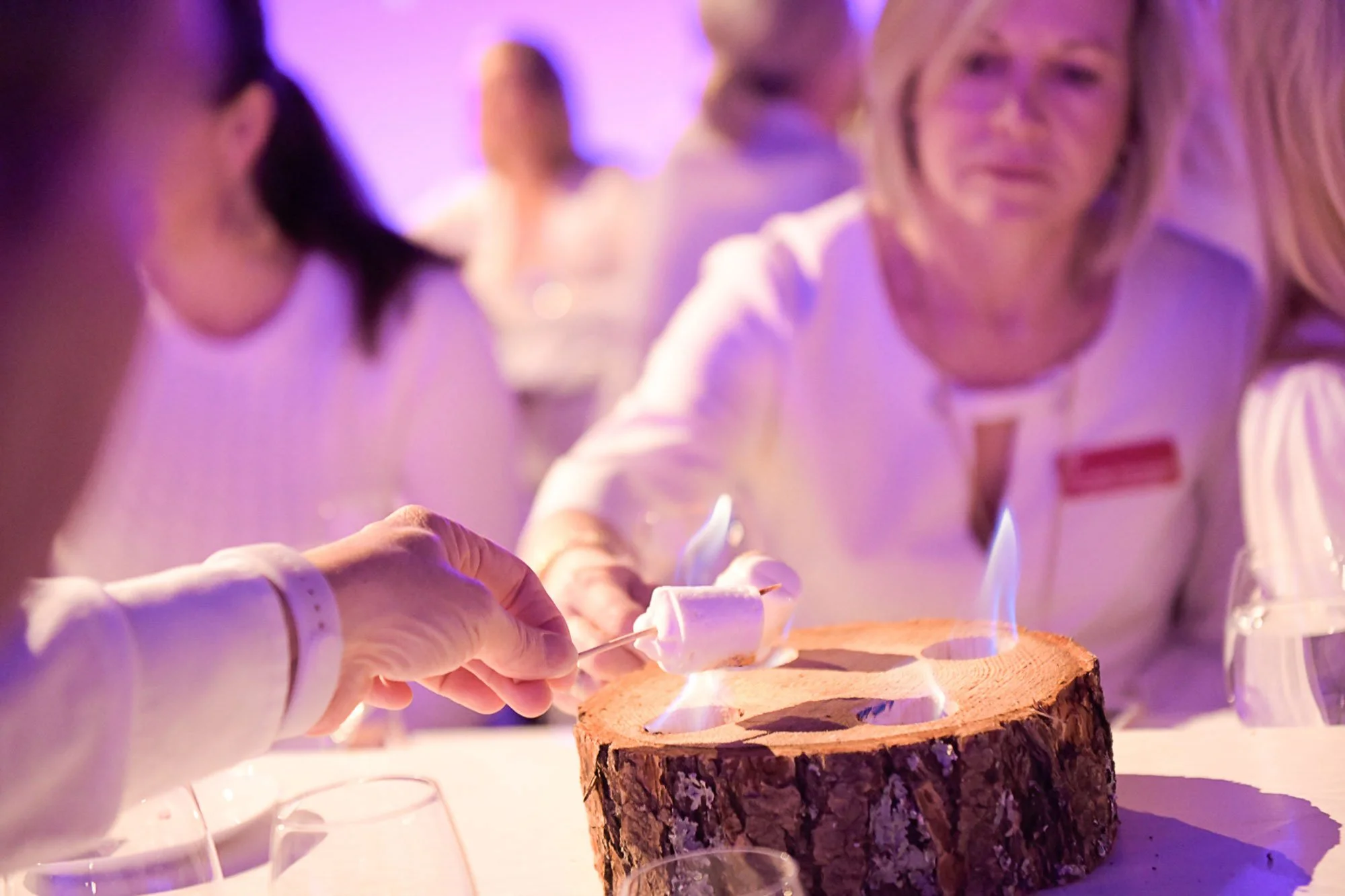 People wearing white shirts preparing to ignite marshmallows on sticks over a fire on a wooden log at a social event with purple lighting.