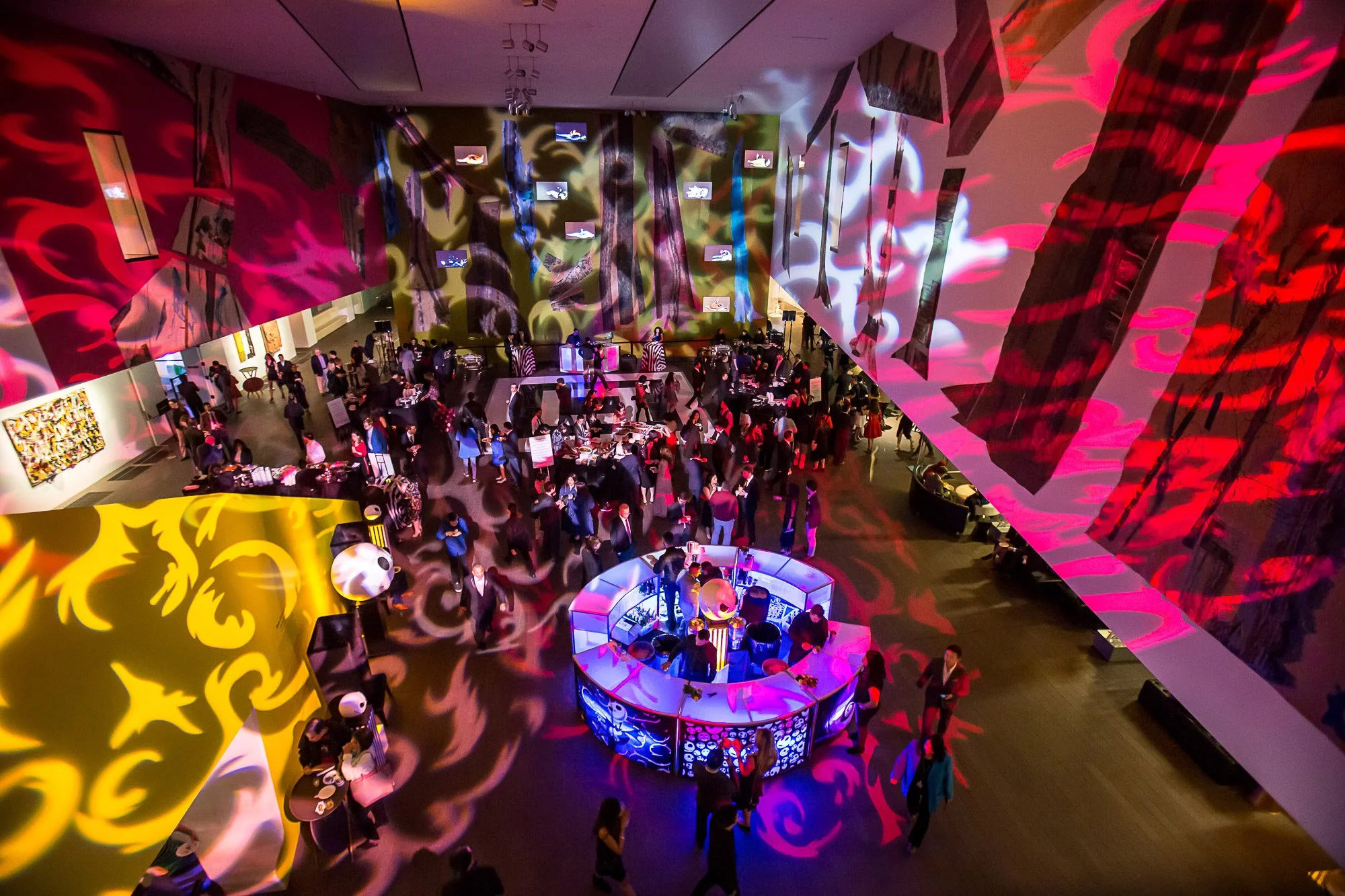 An indoor art exhibition or event with multicolored lighting and projections on the walls and ceiling, featuring people gathered around displays and tables.