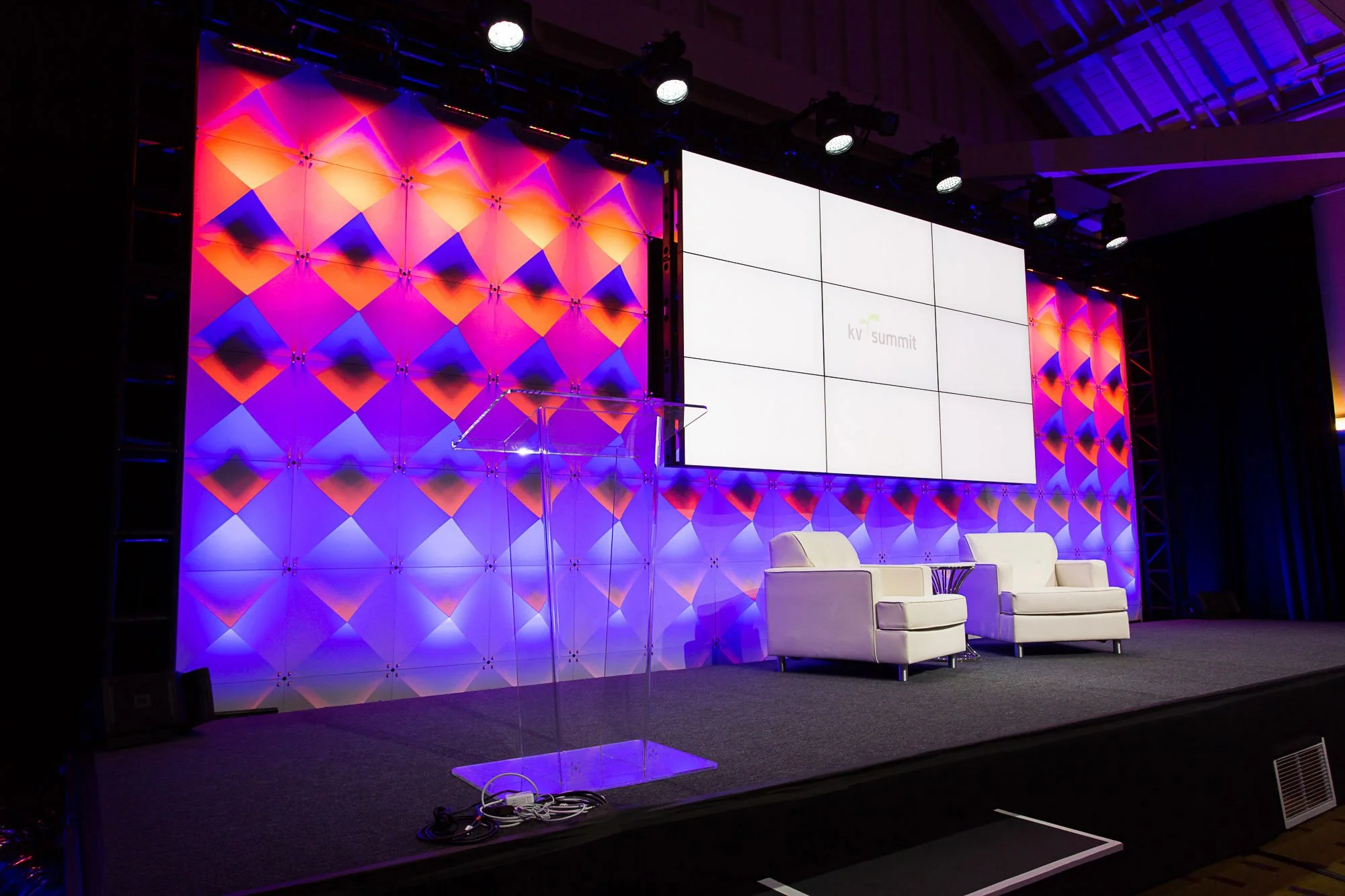 Stage setup for a conference or panel discussion with a colorful geometric background, large screen displaying 'kv summit', and two white chairs with a small table.