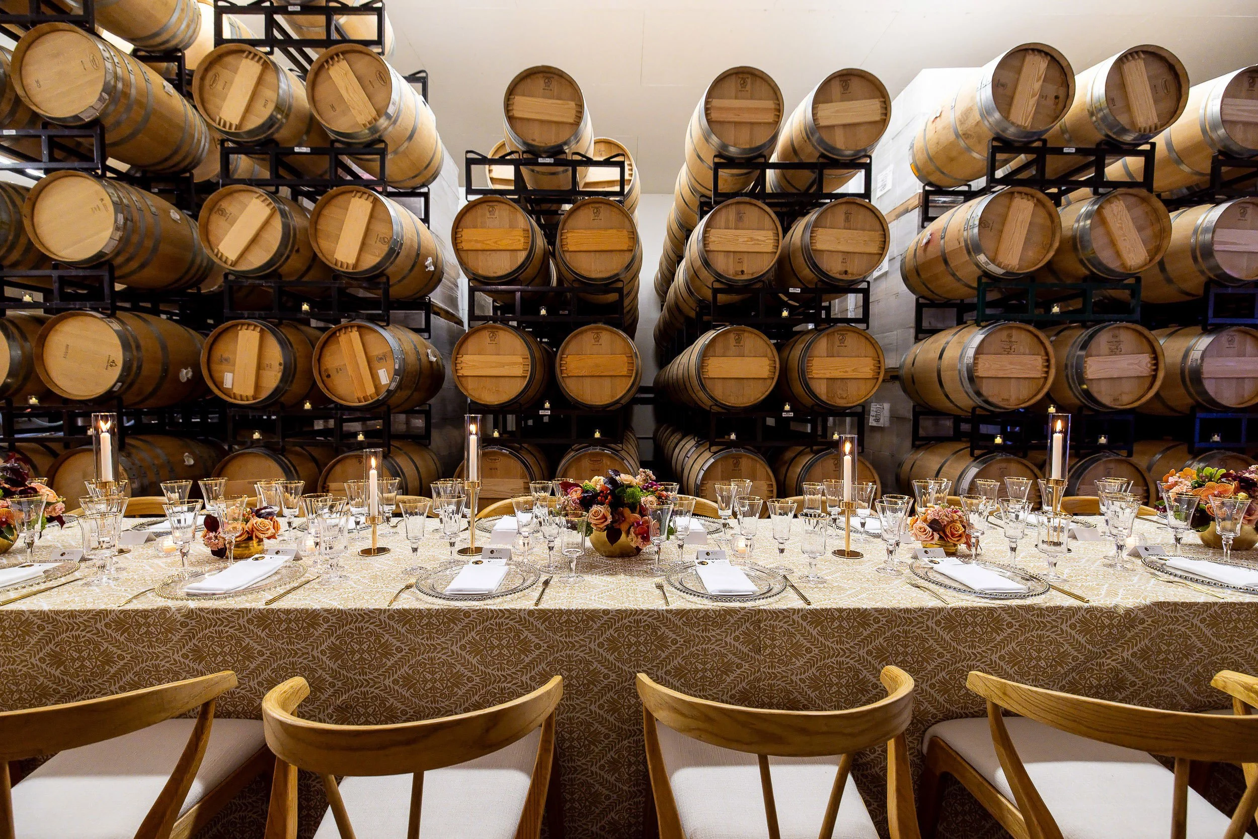 Room with stacked wine barrels on black metal racks and a long banquet table set with glassware, napkins, and floral centerpieces, suggesting a winery or tasting room setting.