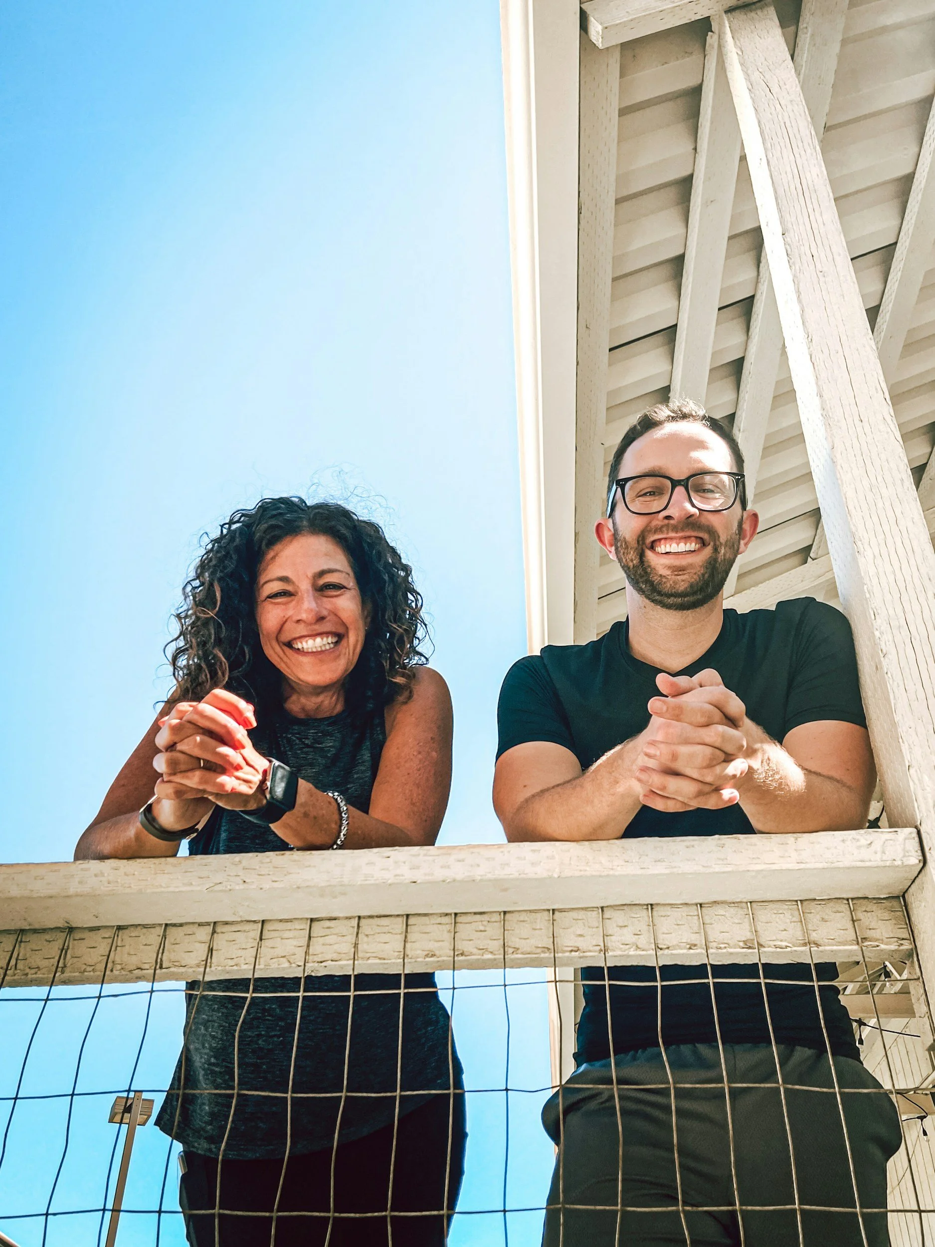 A woman with curly dark hair and a man with glasses and facial hair smiling, leaning on a wooden railing outside against a bright blue sky. They are both holding their hands together near their chins and appear happy.