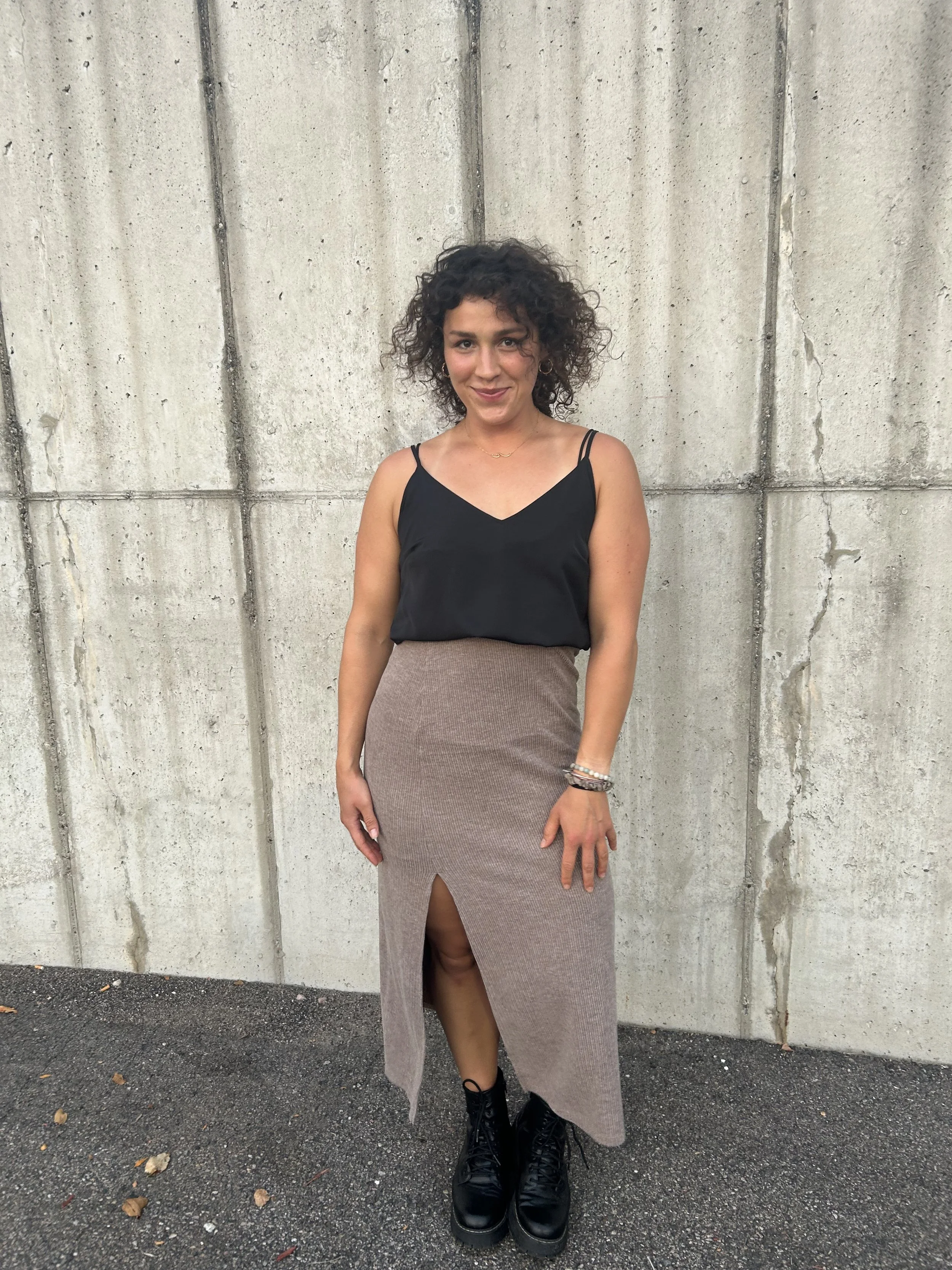 A woman standing outdoors against a concrete wall, wearing a black spaghetti strap top, a beige skirt with a front slit, black boots, and jewelry including bracelets, a necklace, and earrings.