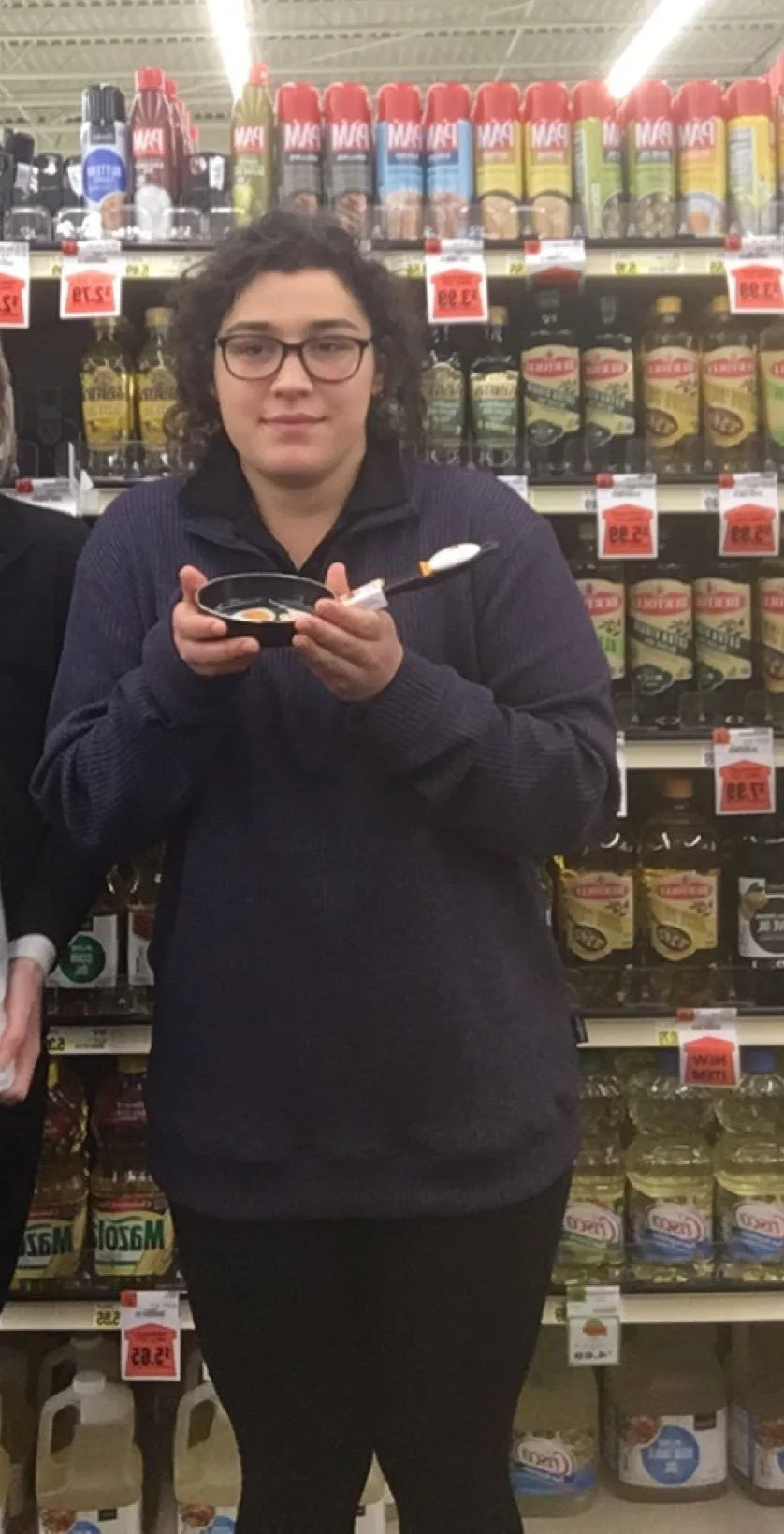 Woman with glasses holding a bowl and spoon in grocery store aisle with condiments on shelves.