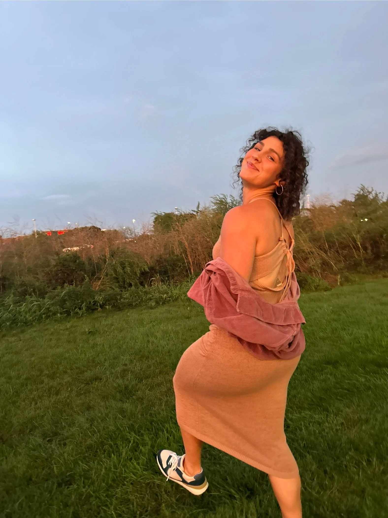 A woman with curly dark hair standing on grass outdoors during sunset, wearing a beige tank top and matching shorts, with a pinkish-brown jacket draped over her shoulders, smiling and looking back at the camera.