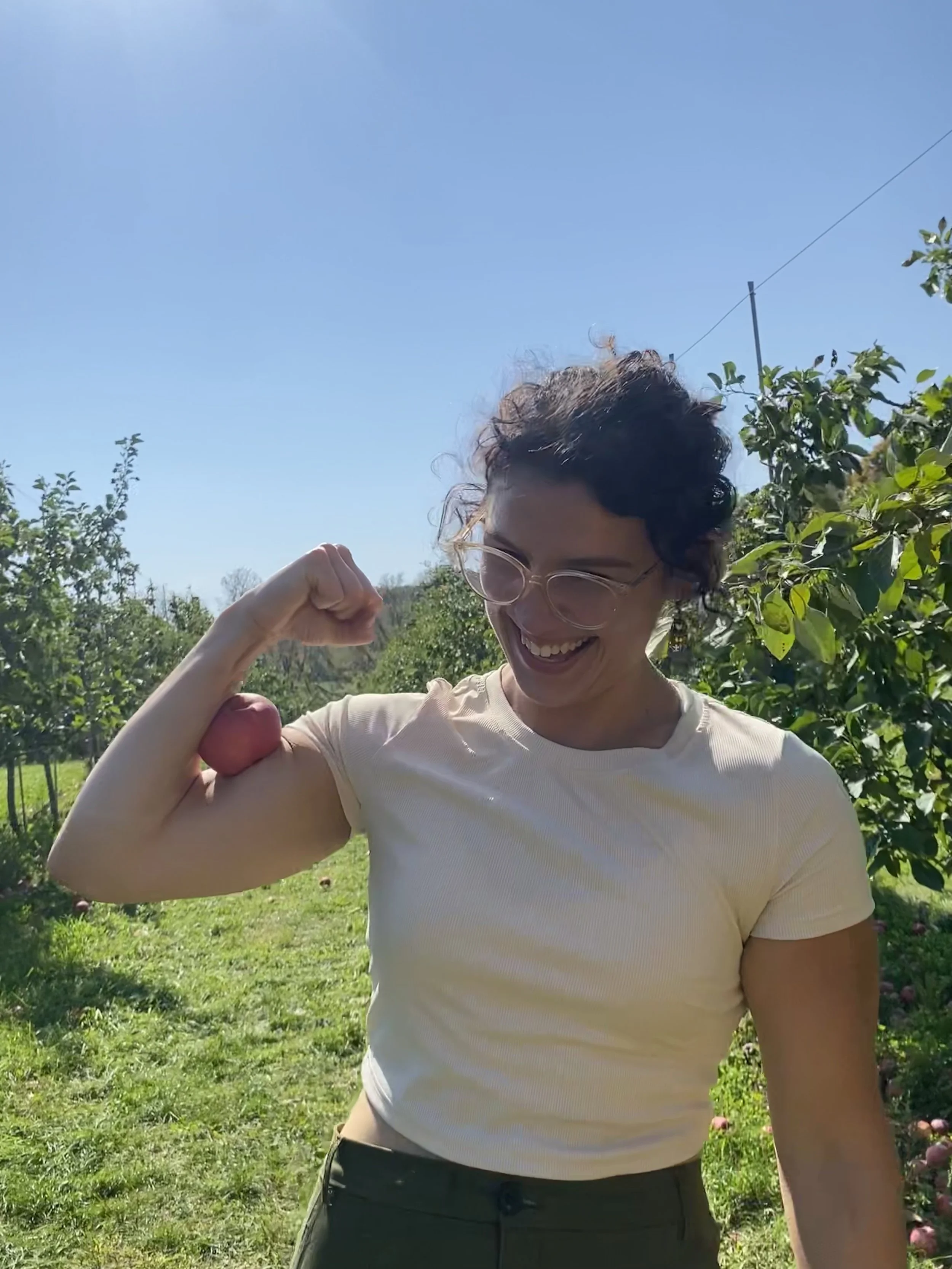 A woman with curly hair and glasses smiling, standing outdoors on a sunny day, flexing her right arm, with trees and blue sky in the background.
