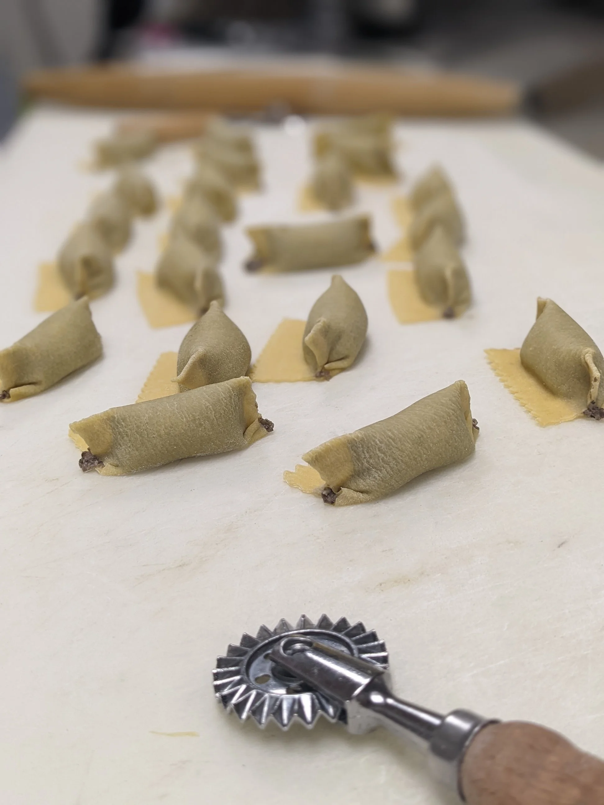 Homemade ravioli on a parchment-lined surface with a pasta cutter tool in the foreground.