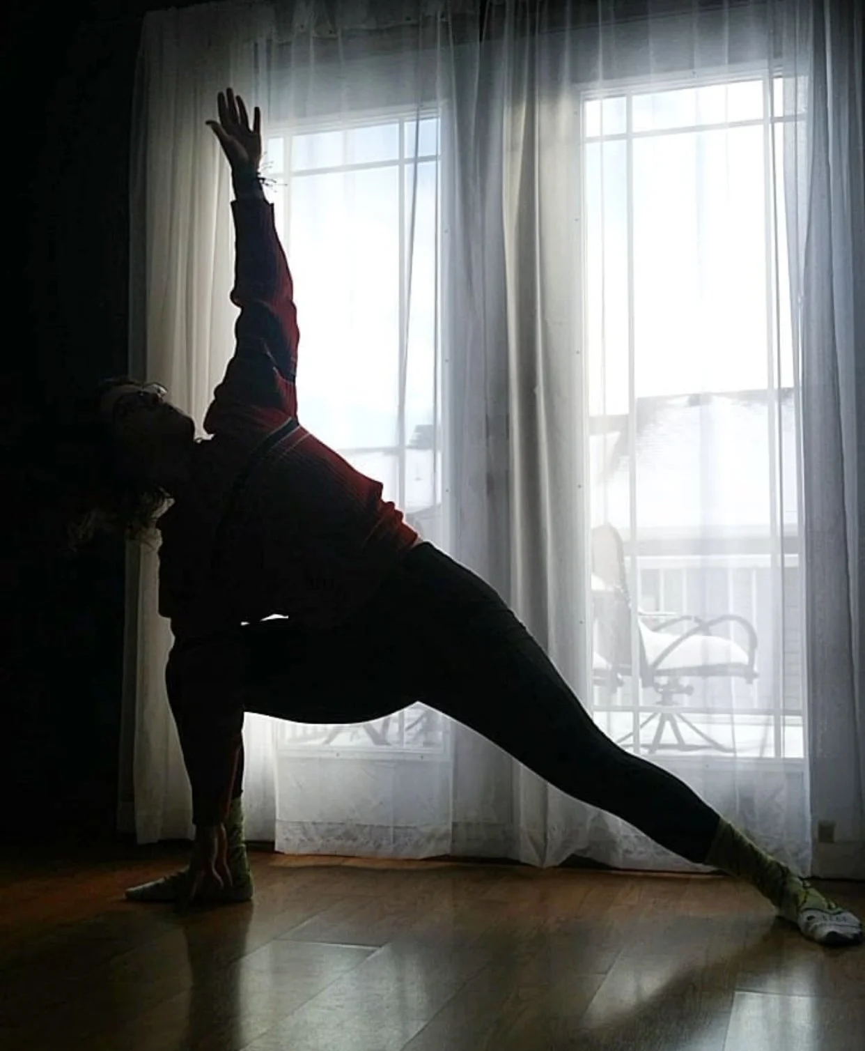 Person doing a side stretch yoga pose in front of a backlit window with sheer curtains, balcony furniture visible outside.