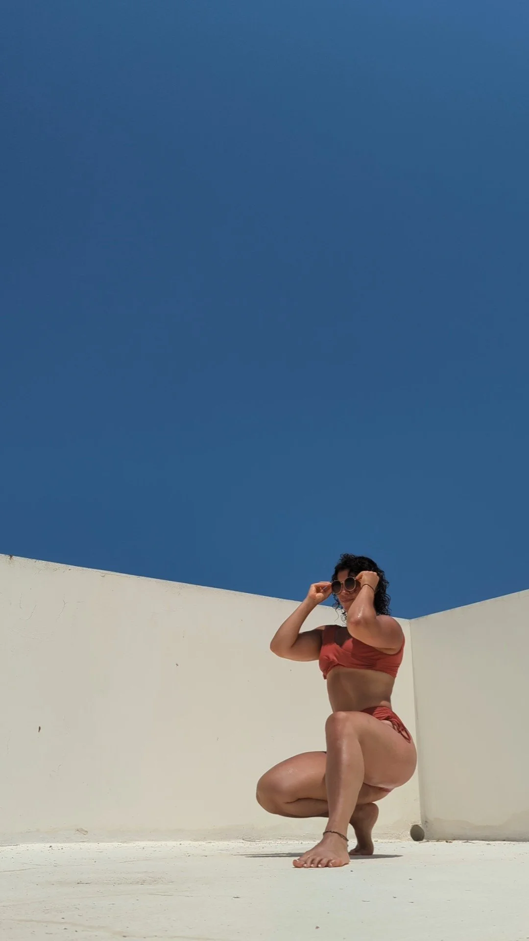 A woman in a red bikini squatting on a white surface, holding sunglasses, with a clear blue sky and white walls in the background.
