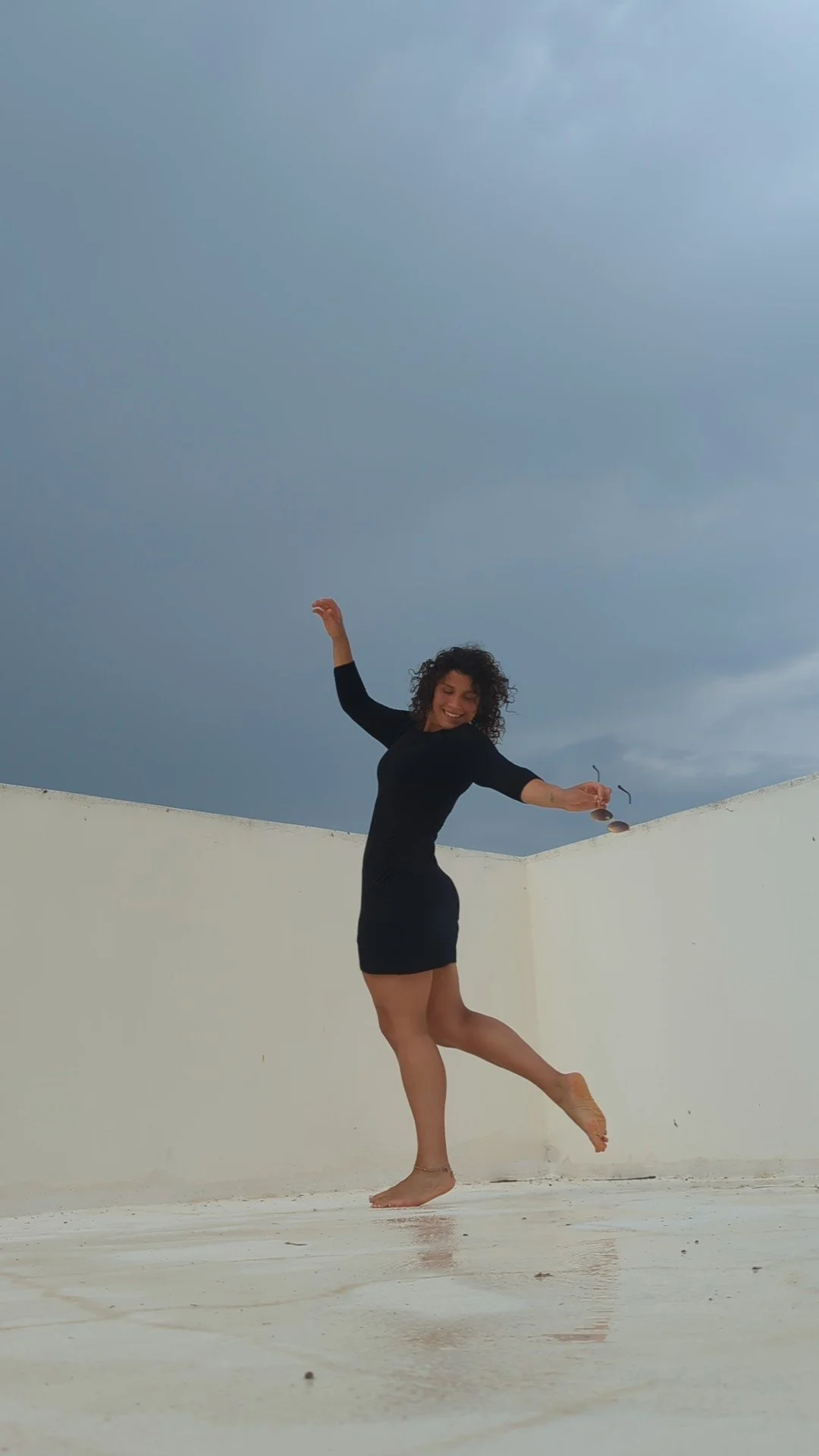 A woman in a black dress is dancing on a white rooftop with a cloudy sky in the background.