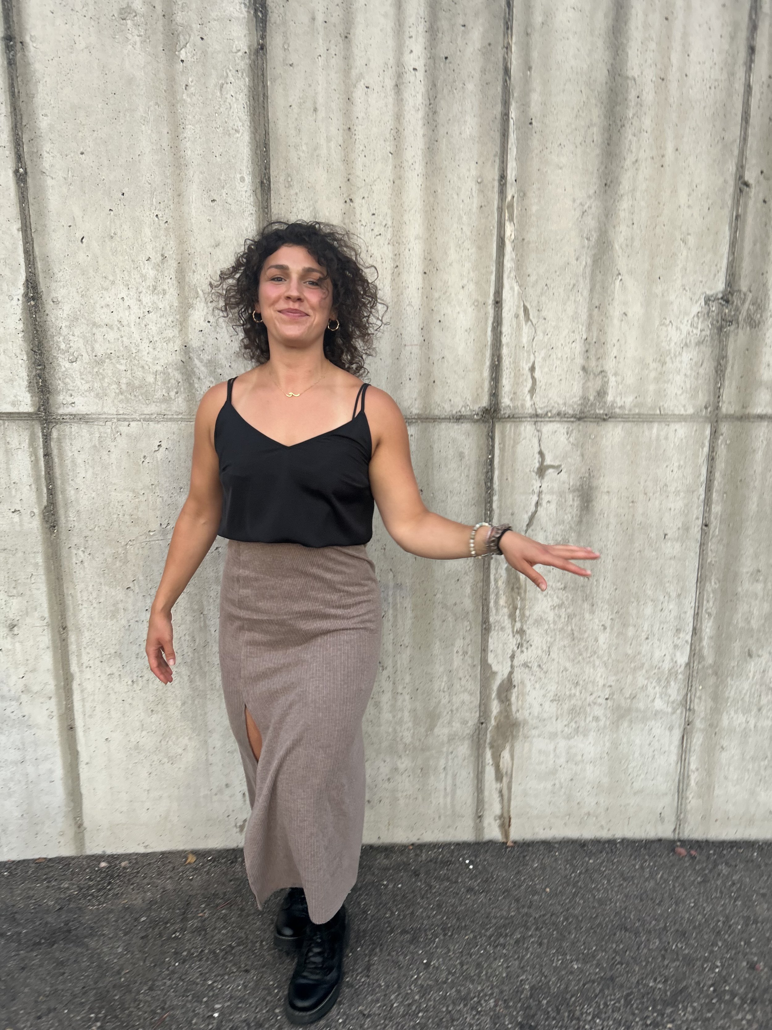 A woman with curly dark hair standing in front of a concrete wall, wearing a black spaghetti strap top, a beige skirt with a slit, black boots, and jewelry including earrings, necklaces, and bracelets.