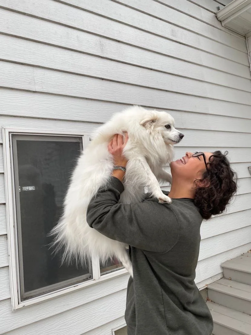 A person with glasses smiling and holding a fluffy white dog outside next to a house with white siding.