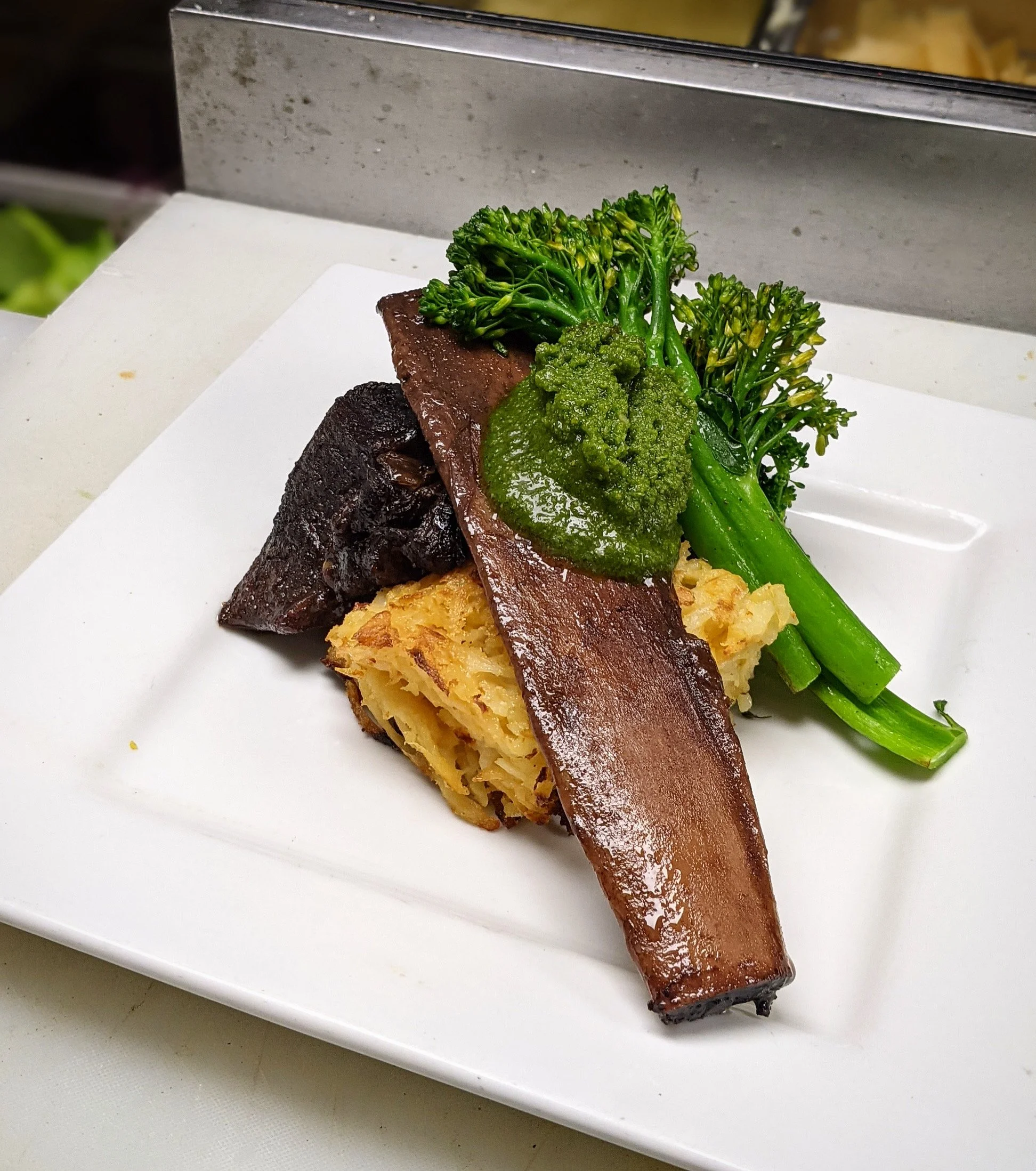 A plated dish featuring braised beef, slices of seasoned green broccolini, and a piece of potato casserole on a white rectangular plate.