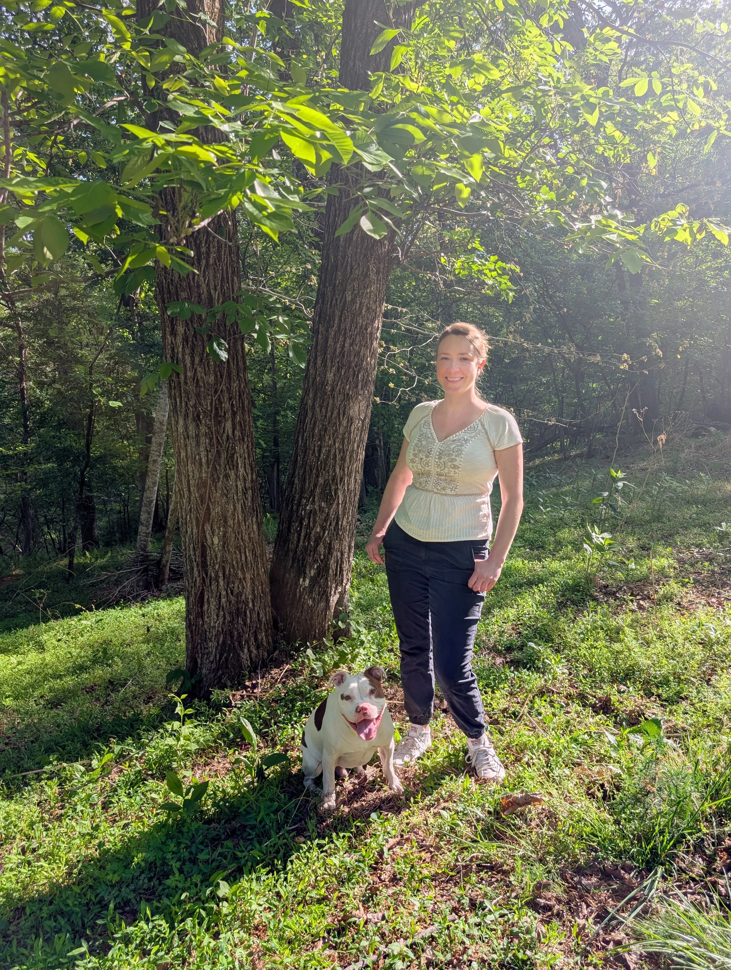 A woman with light brown hair smiling, standing outdoors in a wooded area with green leaves and tall trees, wearing a light-colored embroidered top, dark pants, and white sneakers, accompanied by a white and black dog sitting on the grass in front of her, with sunlight filtering through the trees.