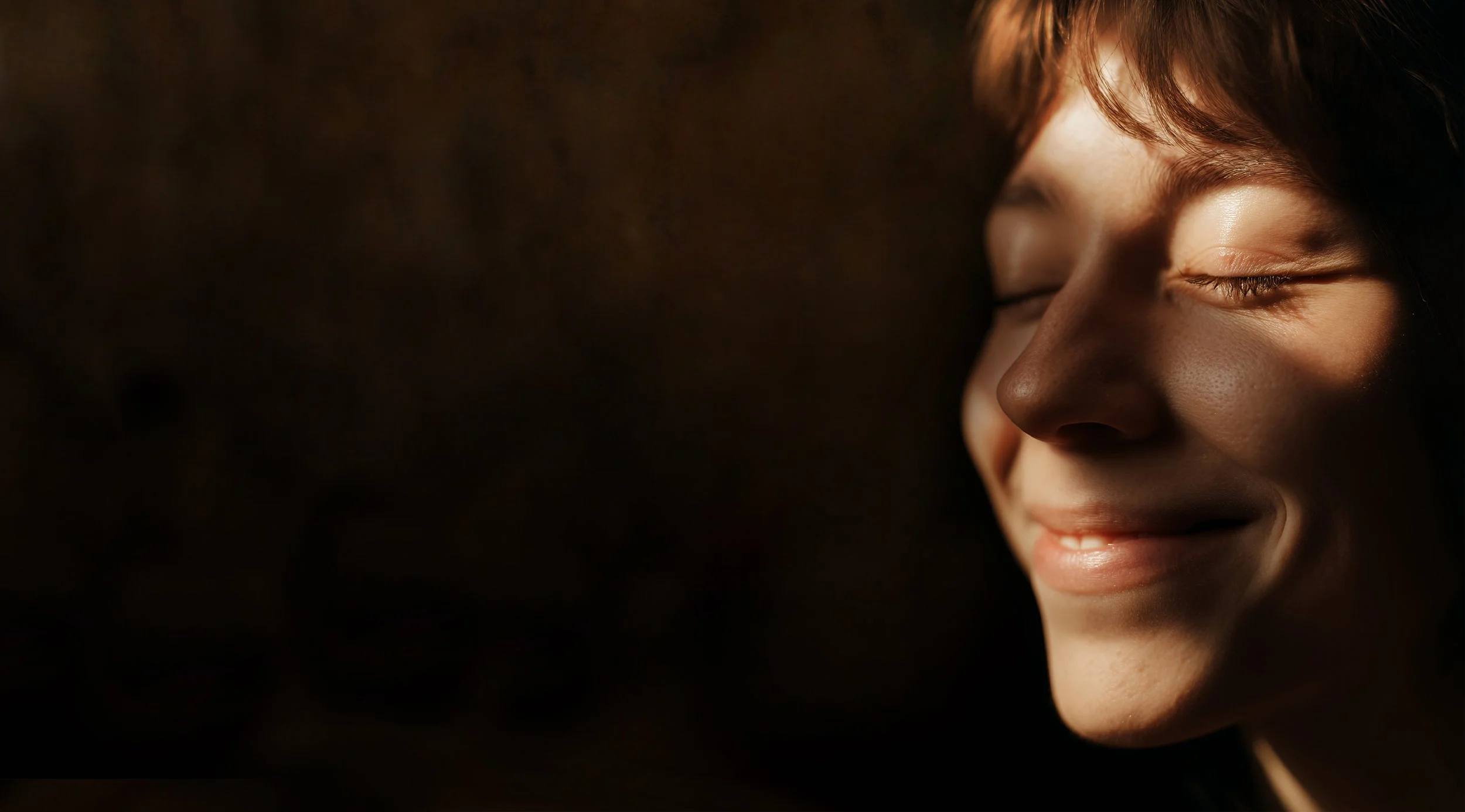 Close-up of a young woman's face with closed eyes, smiling softly, with warm lighting creating a peaceful mood.