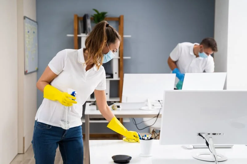 Two people in a modern office wearing masks and gloves, cleaning desks and computer monitors.