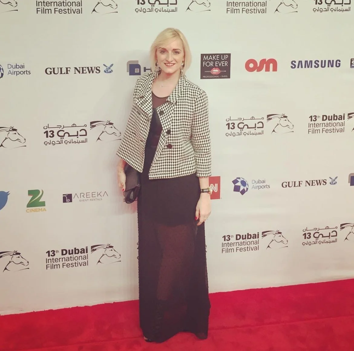 A woman with blonde hair wearing a black and white checkered jacket and a long black skirt standing on a red carpet at the Dubai International Film Festival.