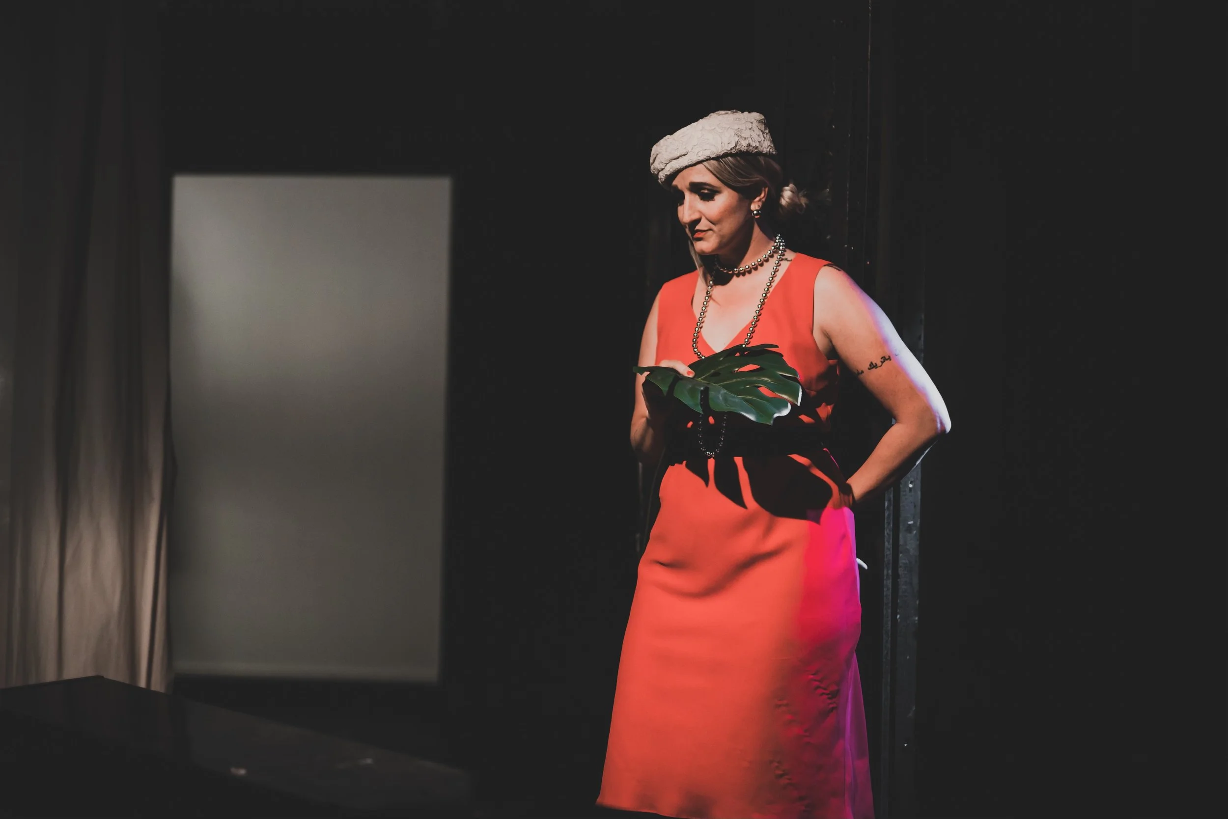 A woman standing in front of a dark background, wearing a red dress, a white hat, and holding a large green leaf, looking at a computer screen.