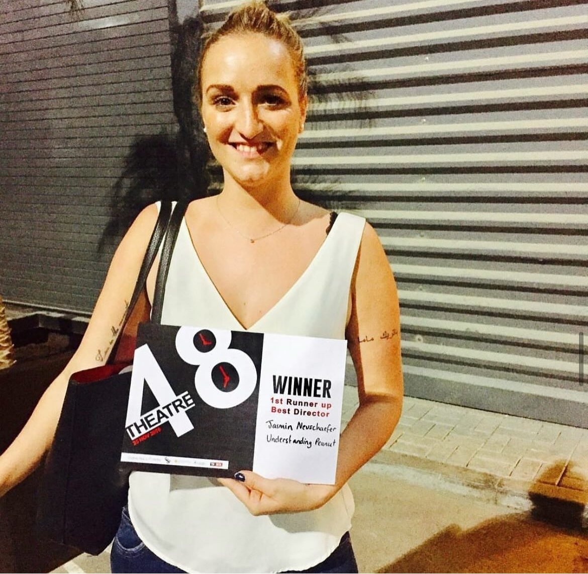 Woman smiling holding an award certificate and a tote bag in front of a metal wall.
